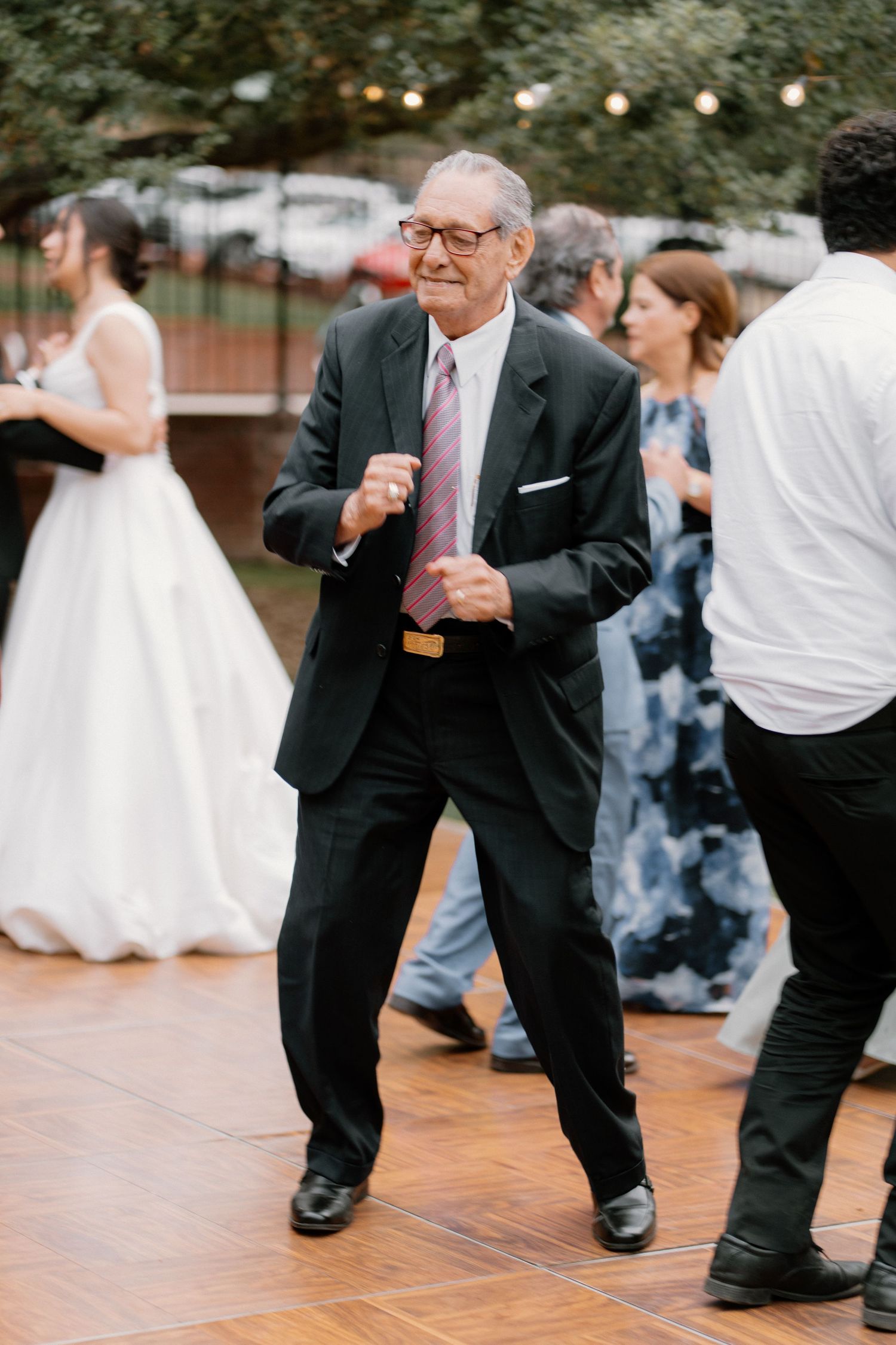 Elderly man in black suit dancing joyfully at an outdoor wedding reception on a wooden dance floor.