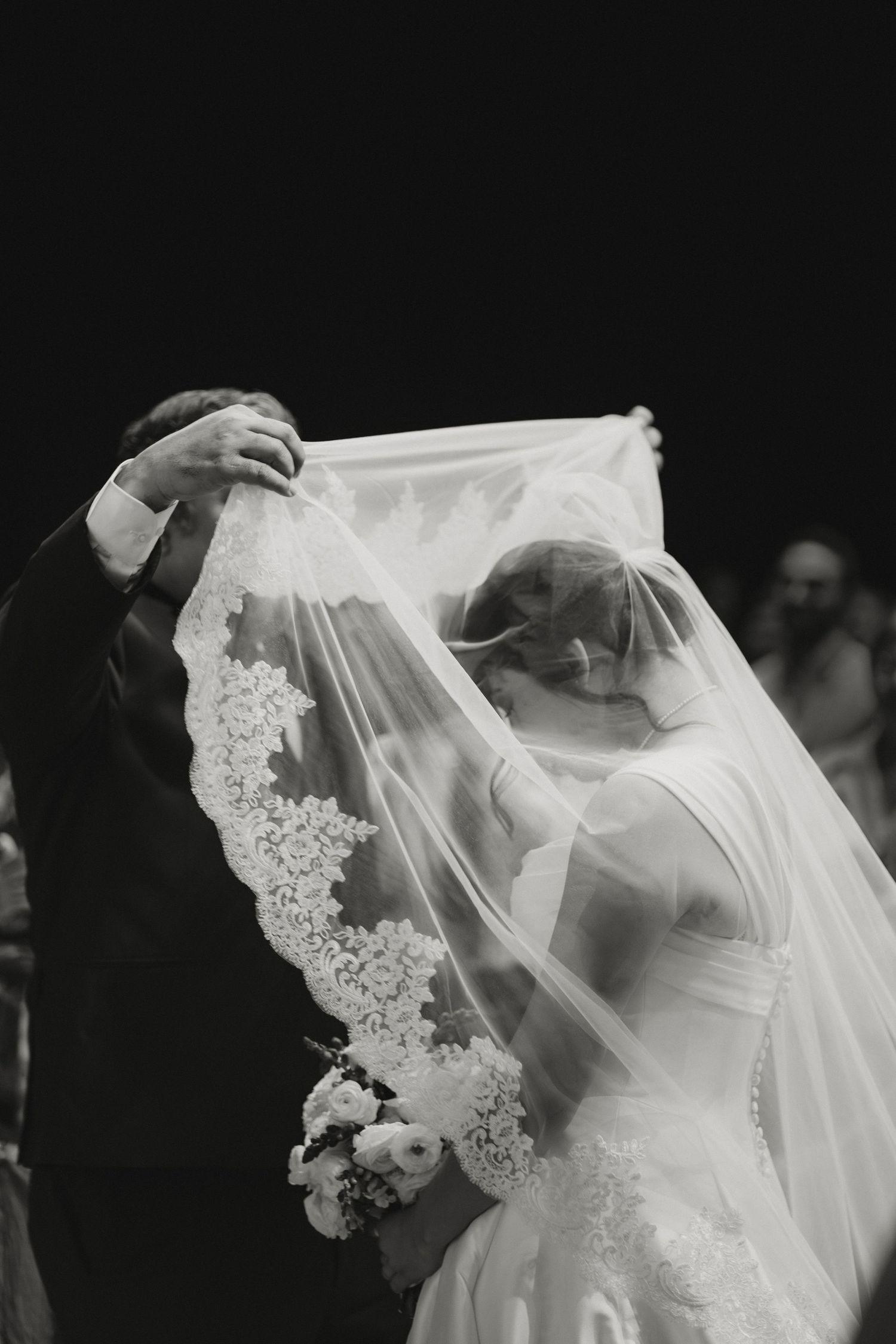 Black and white photo of groom lifting lace veil to kiss bride during ceremony.