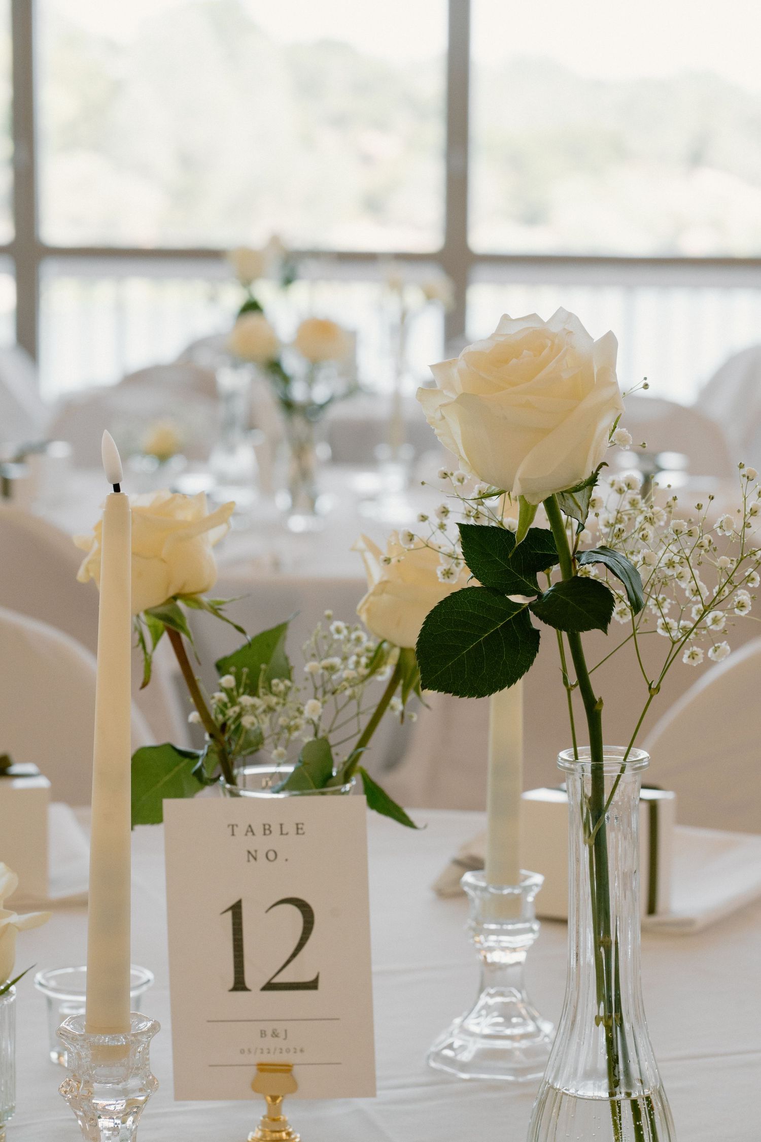 Elegant wedding table centerpiece with white roses, baby breath, taper candles, and table number 12 card.