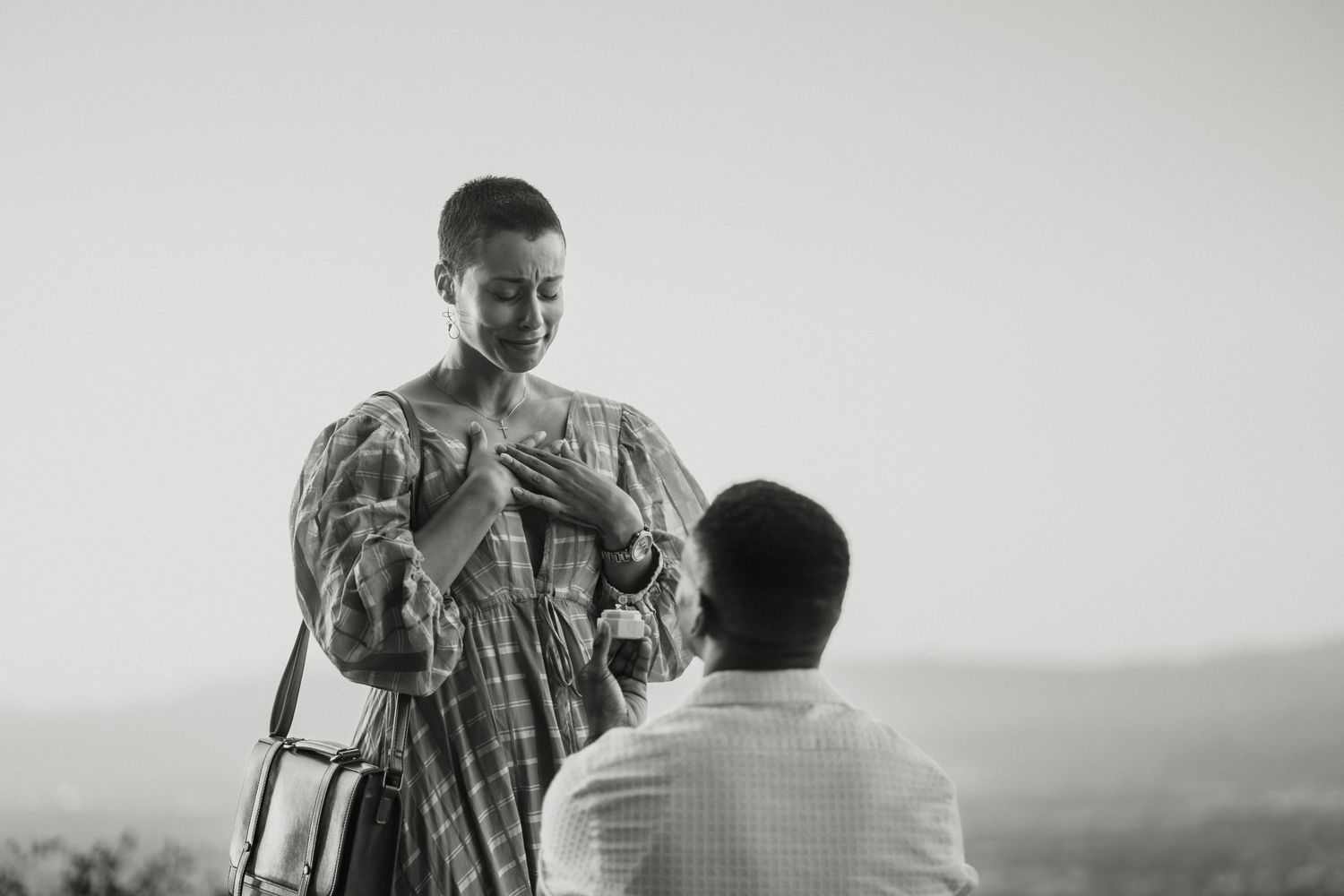 Black and white photo of a man proposing to a woman outdoors in a scenic setting.