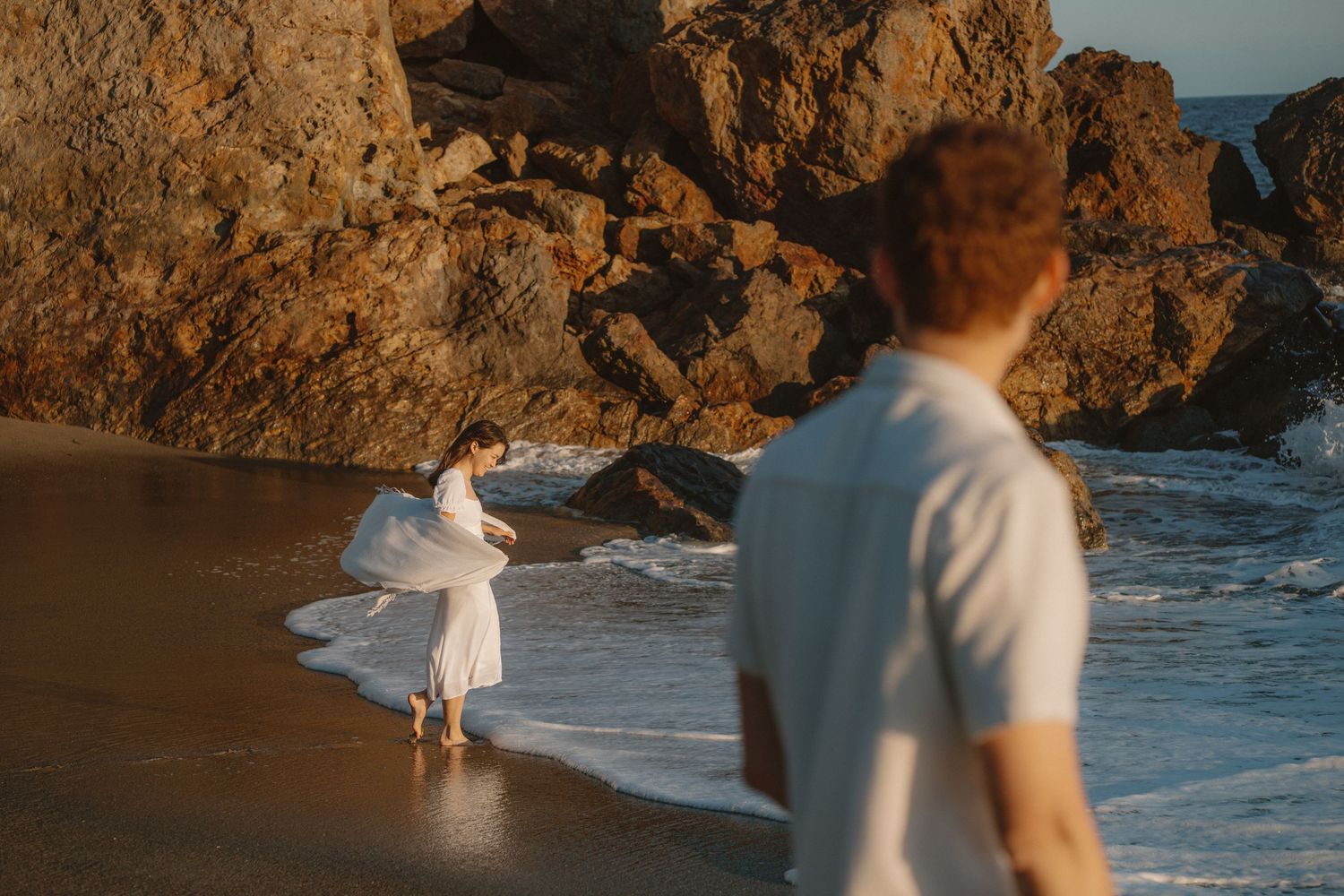 Couple at golden hour on rocky beach, woman in white dress dancing in ocean waves while man watches in foreground.