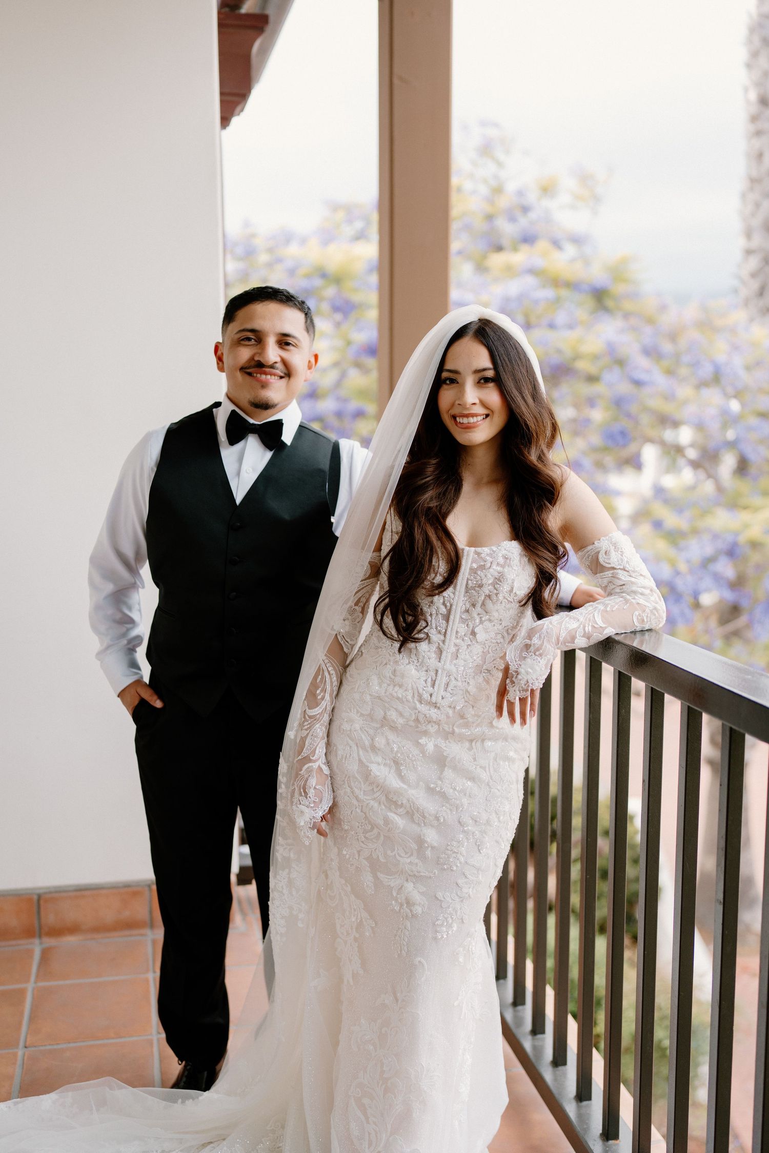 Smiling bride and groom pose together on a balcony, she in lace gown and veil, he in black vest and bow tie.
