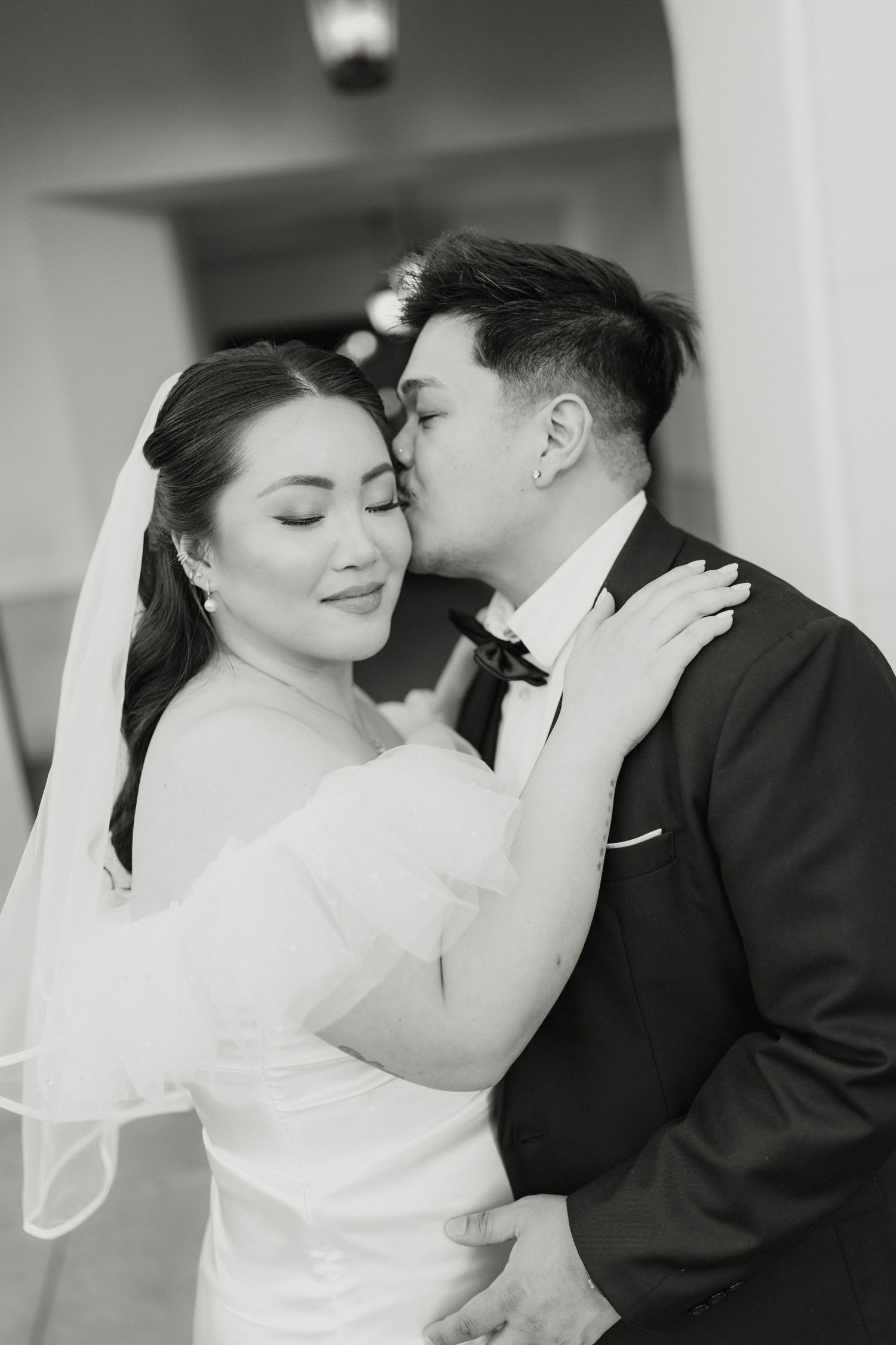 Groom tenderly kisses bride on the cheek in a black and white wedding portrait.
