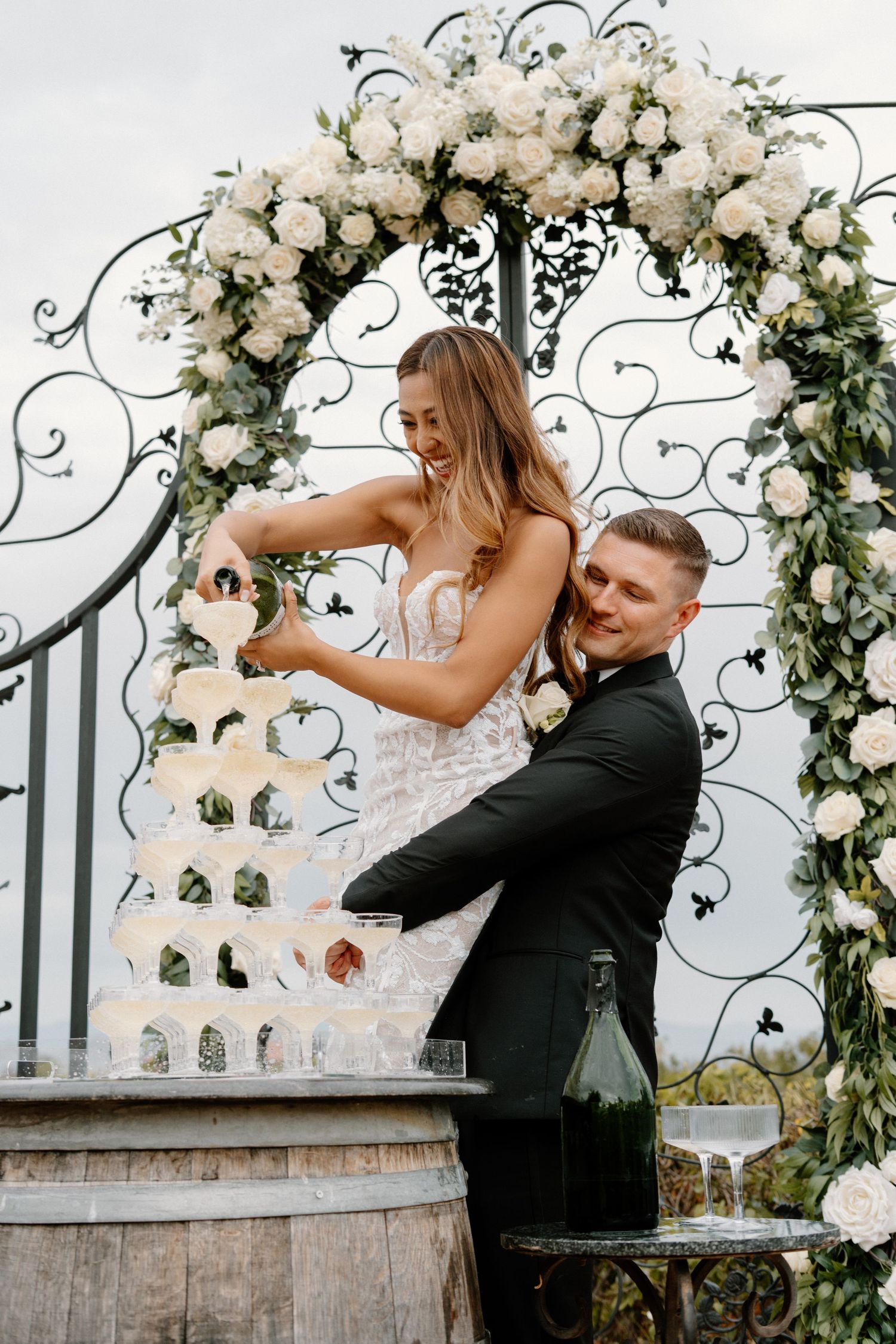 Bride and groom pour champagne tower together beneath ornate floral arch at outdoor wedding reception.