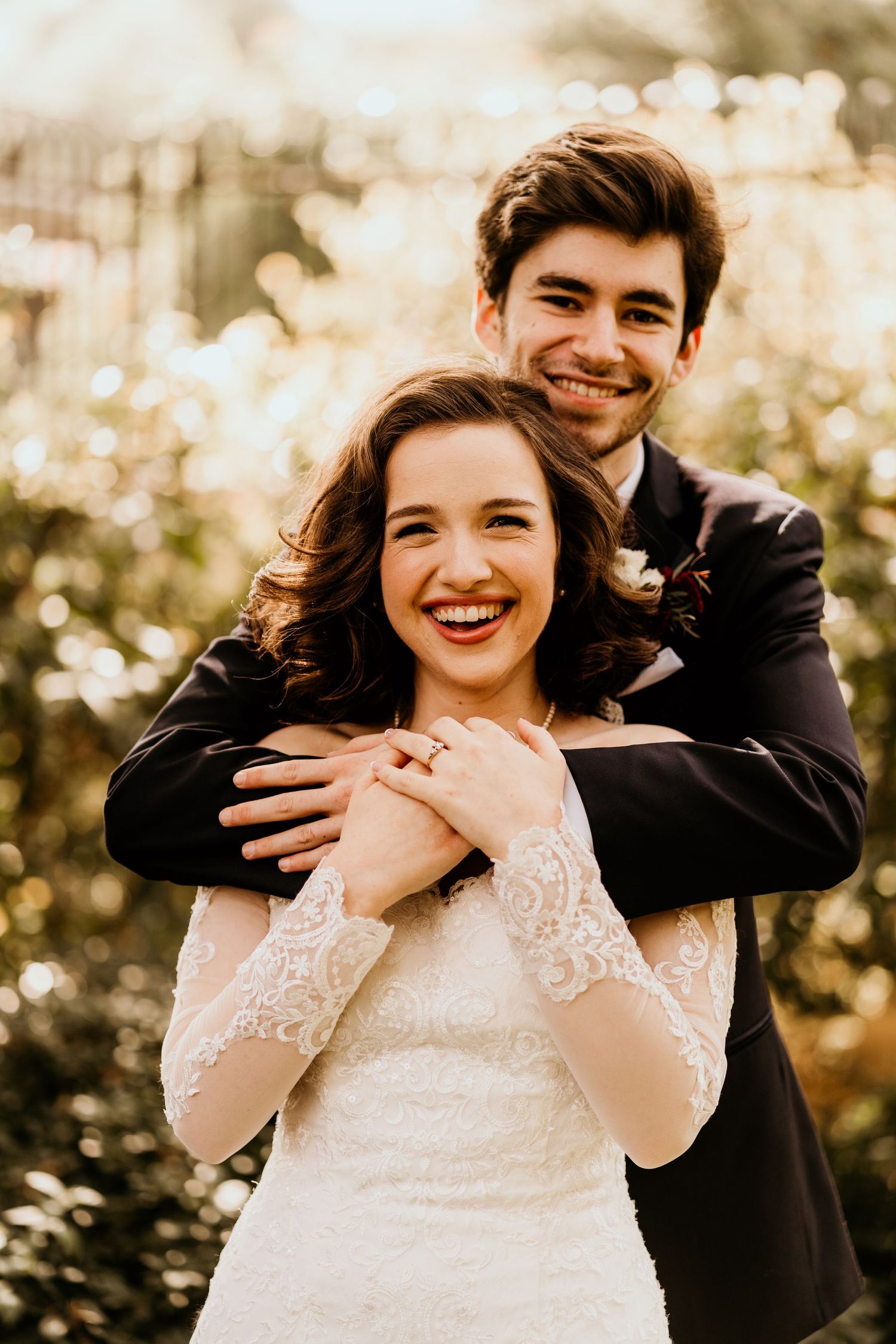 Smiling bride and groom embracing outdoors with bokeh lights, groom hugging bride from behind in wedding attire.
