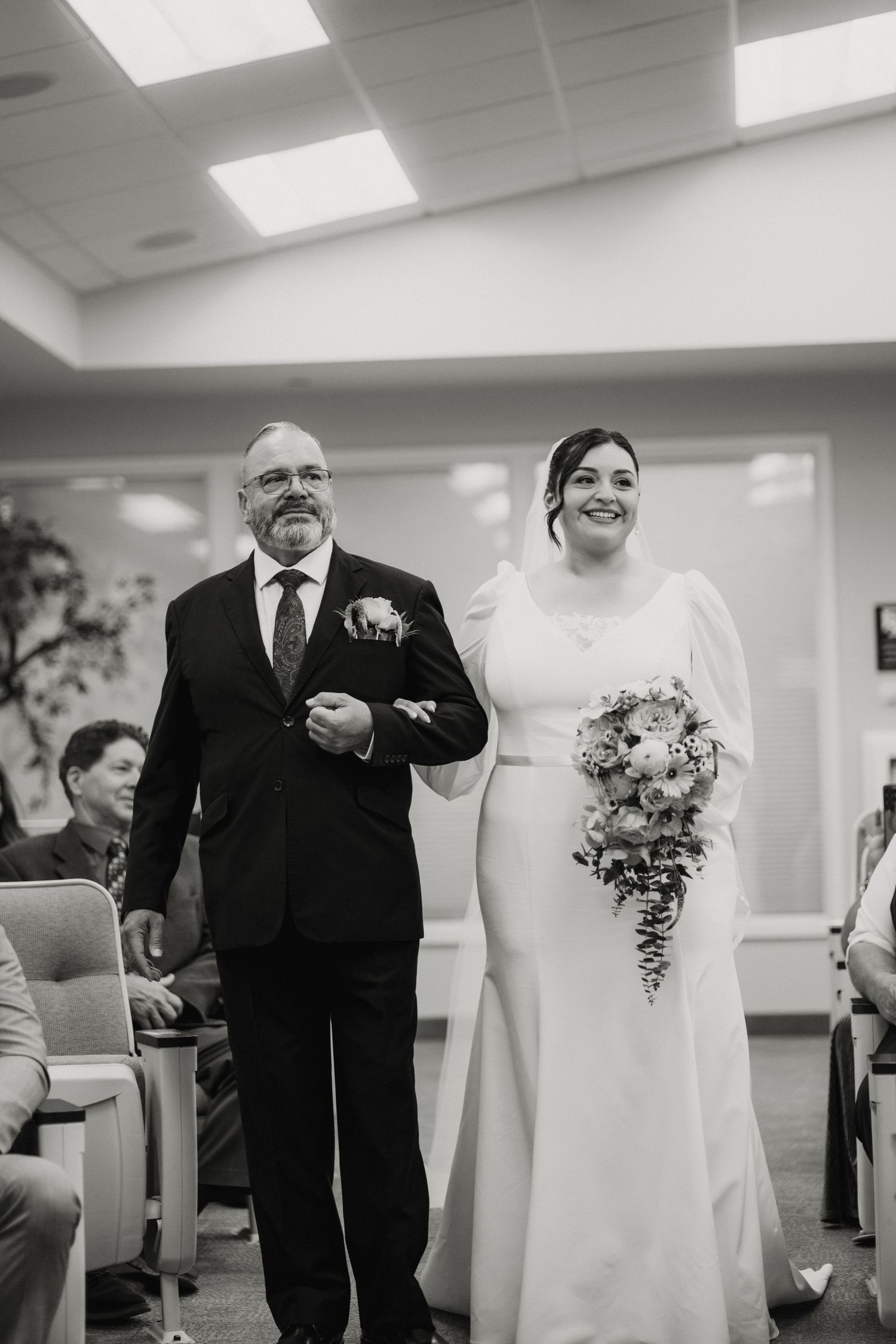 Bride in white long-sleeve gown walks down aisle with escort holding bouquet in black and white photo.