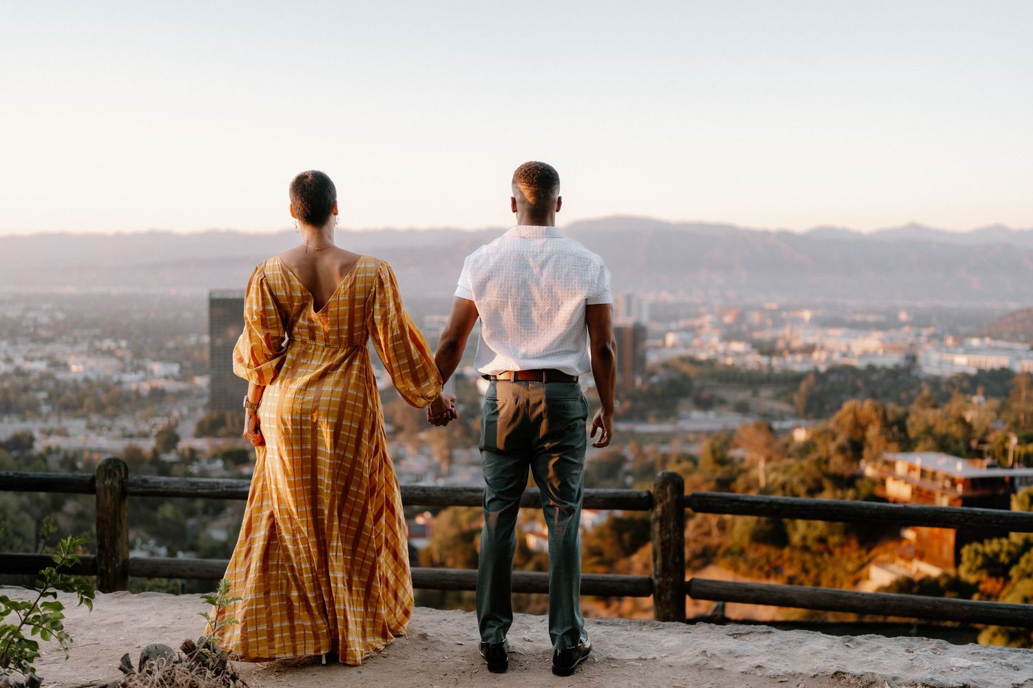 Couple holding hands overlooking a city skyline at sunset from a scenic overlook point.