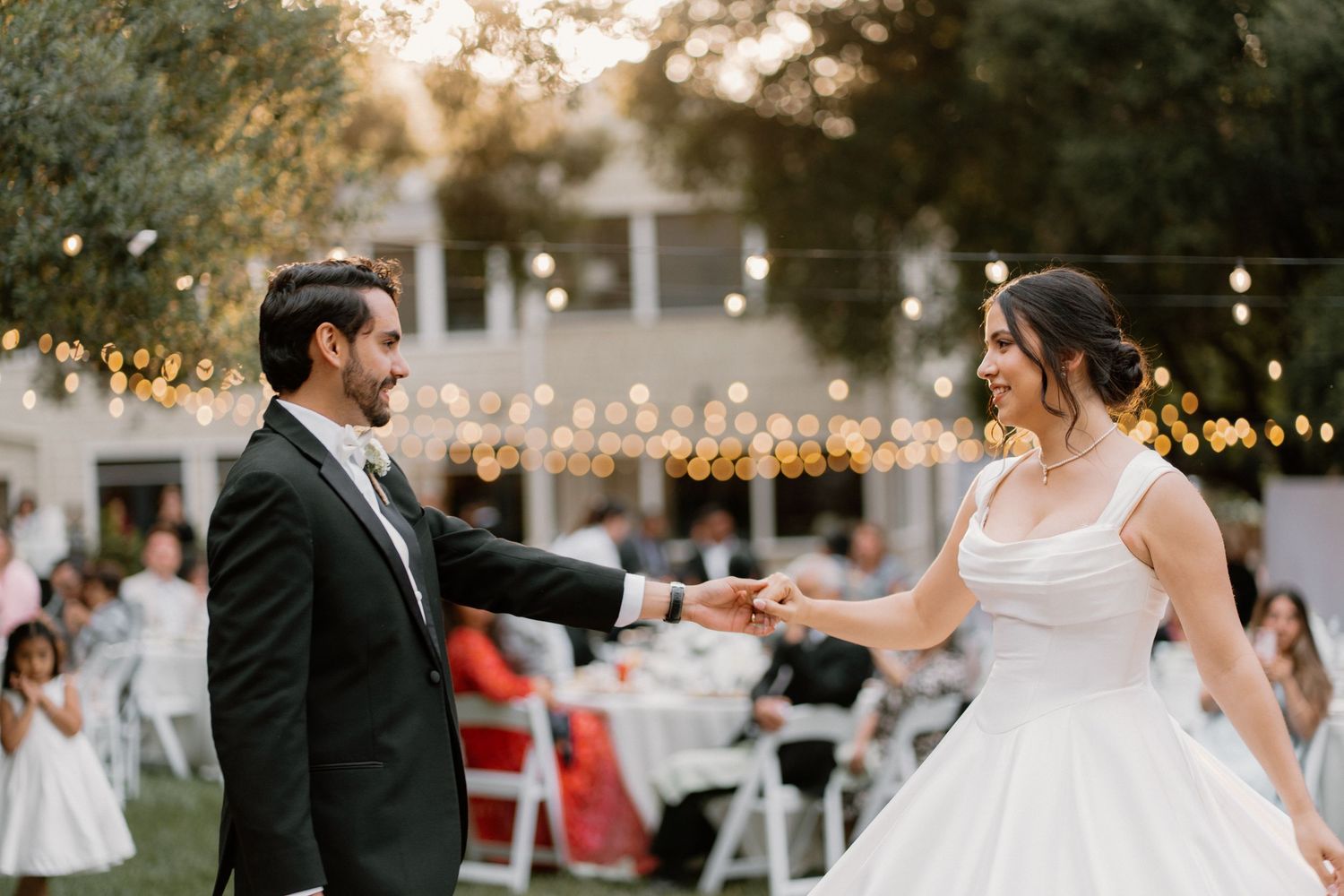 Groom spins smiling bride on outdoor dance floor at golden hour wedding reception with string lights overhead.