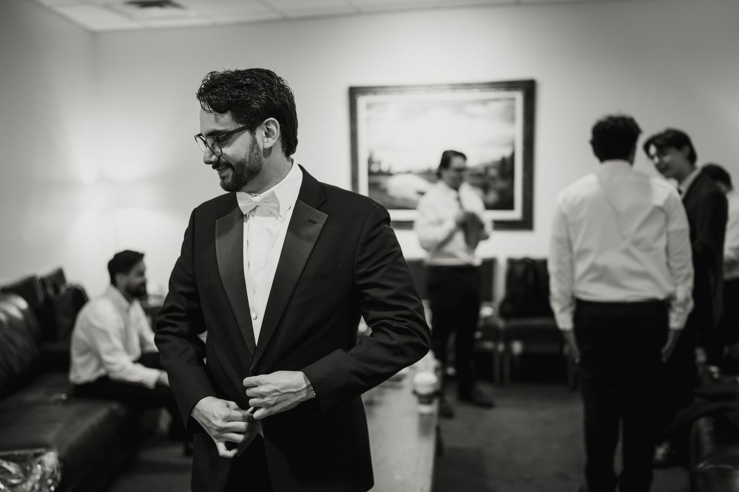 Groom in black tuxedo adjusts cufflinks while groomsmen prepare in background before wedding ceremony.