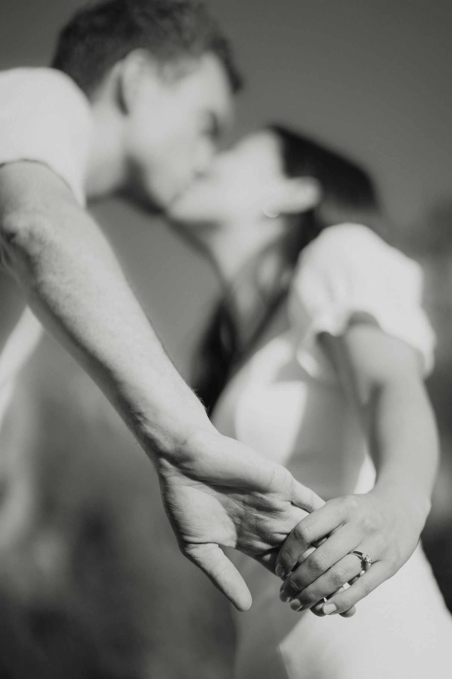 Black and white photo of couple kissing while holding hands, showcasing an engagement ring.