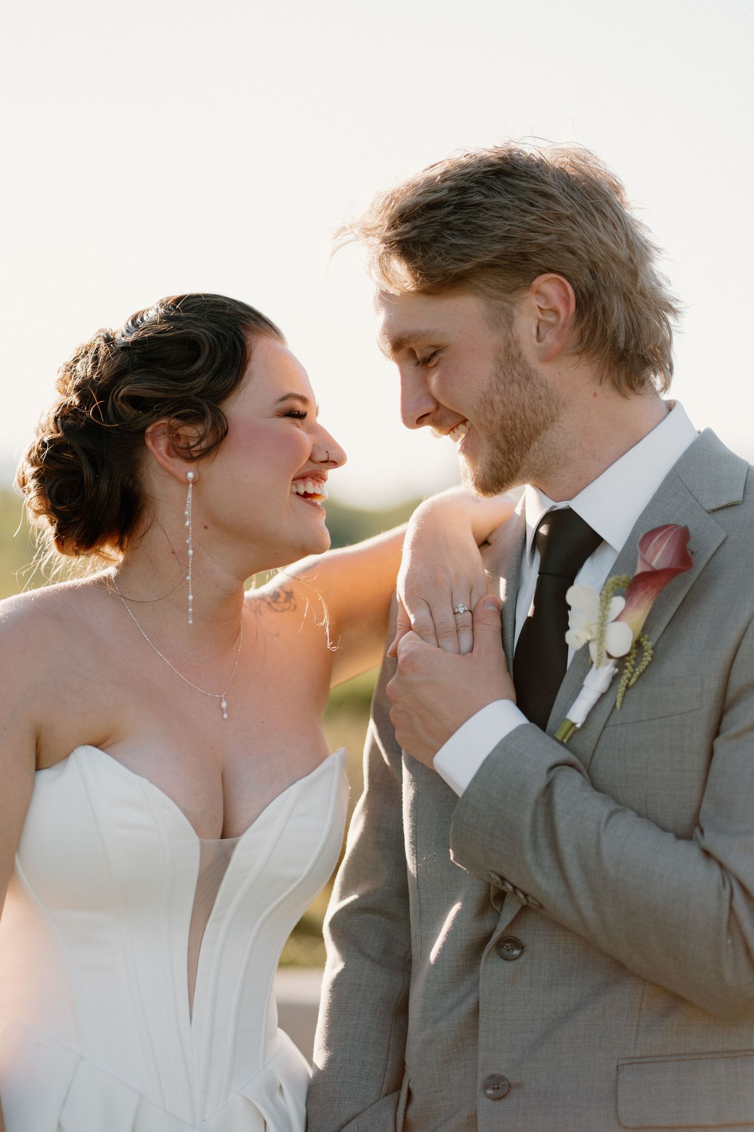 Smiling bride and groom share a tender moment at golden hour, she in a strapless gown, he in a gray suit with boutonniere.