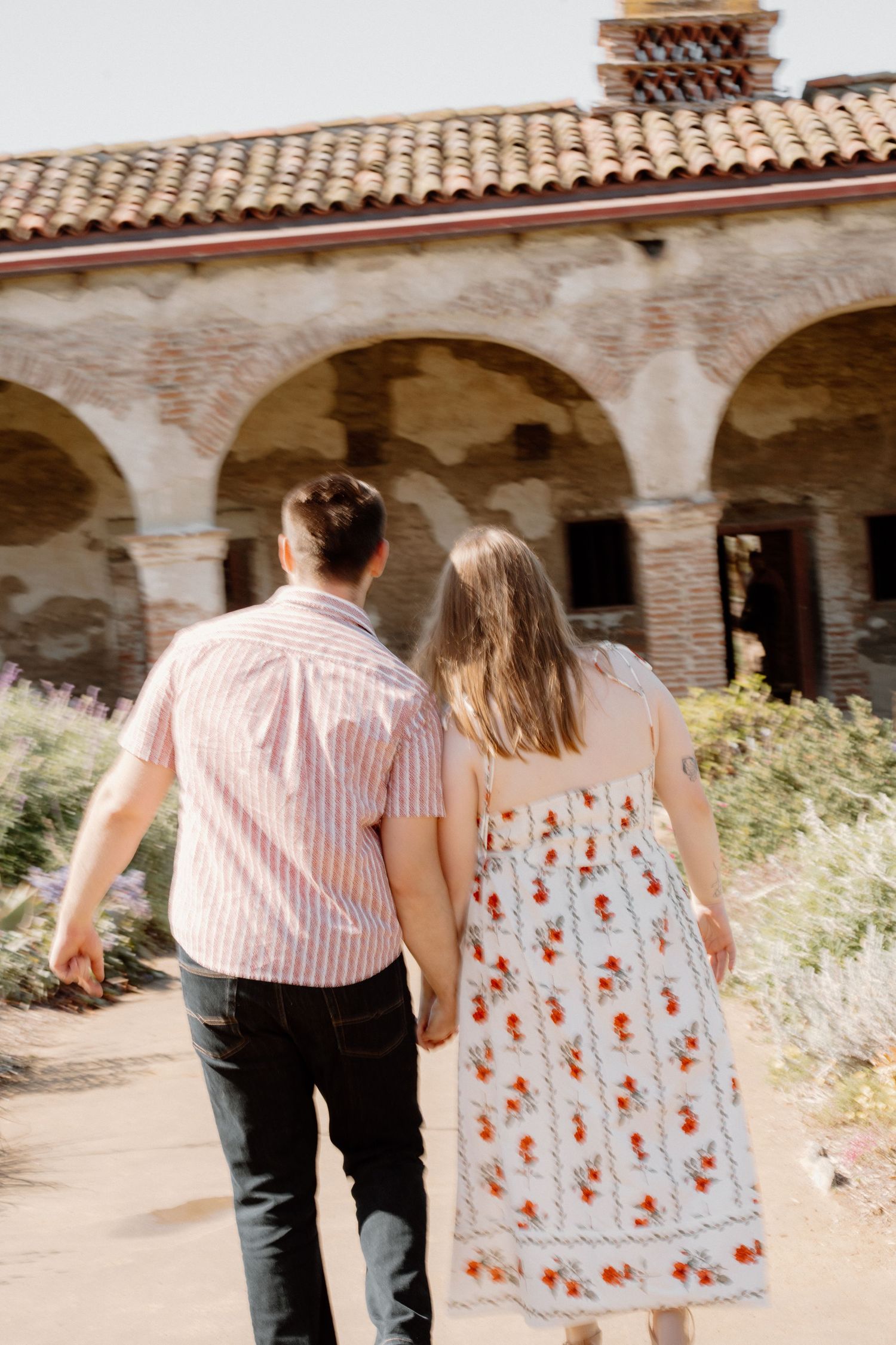 Couple holding hands walking toward a historic Spanish mission with arched colonnade and terracotta roof tiles.