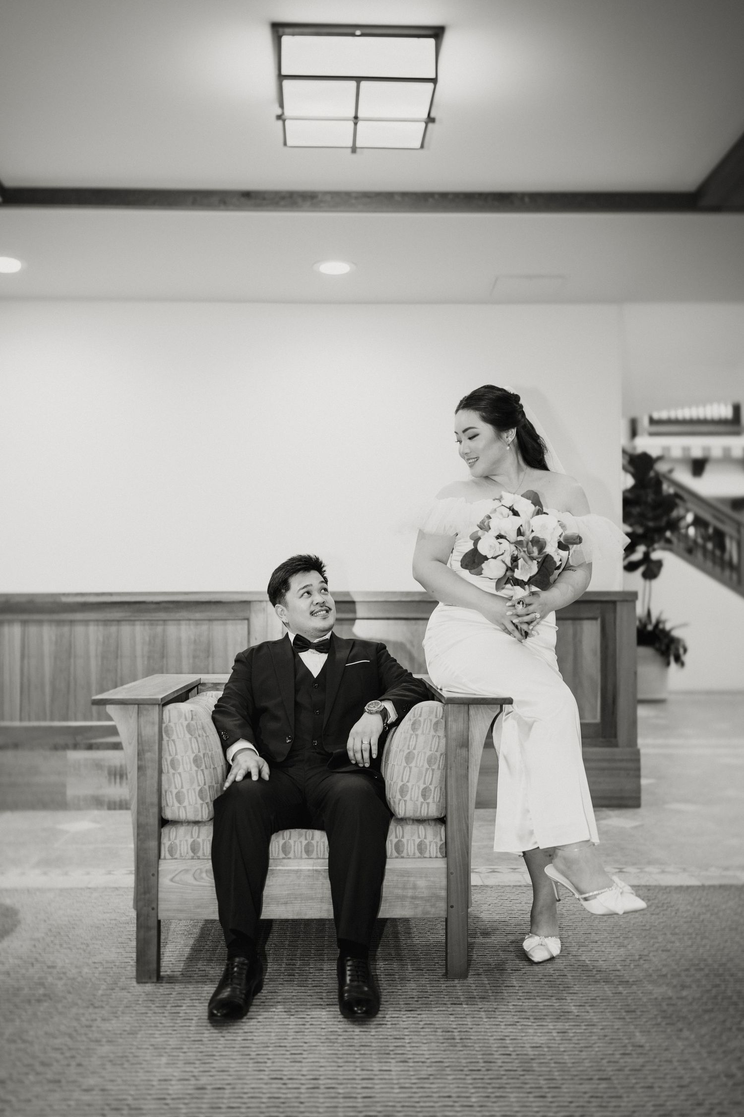 Black and white photo of bride and groom relaxing on a bench, smiling at each other in a lobby.