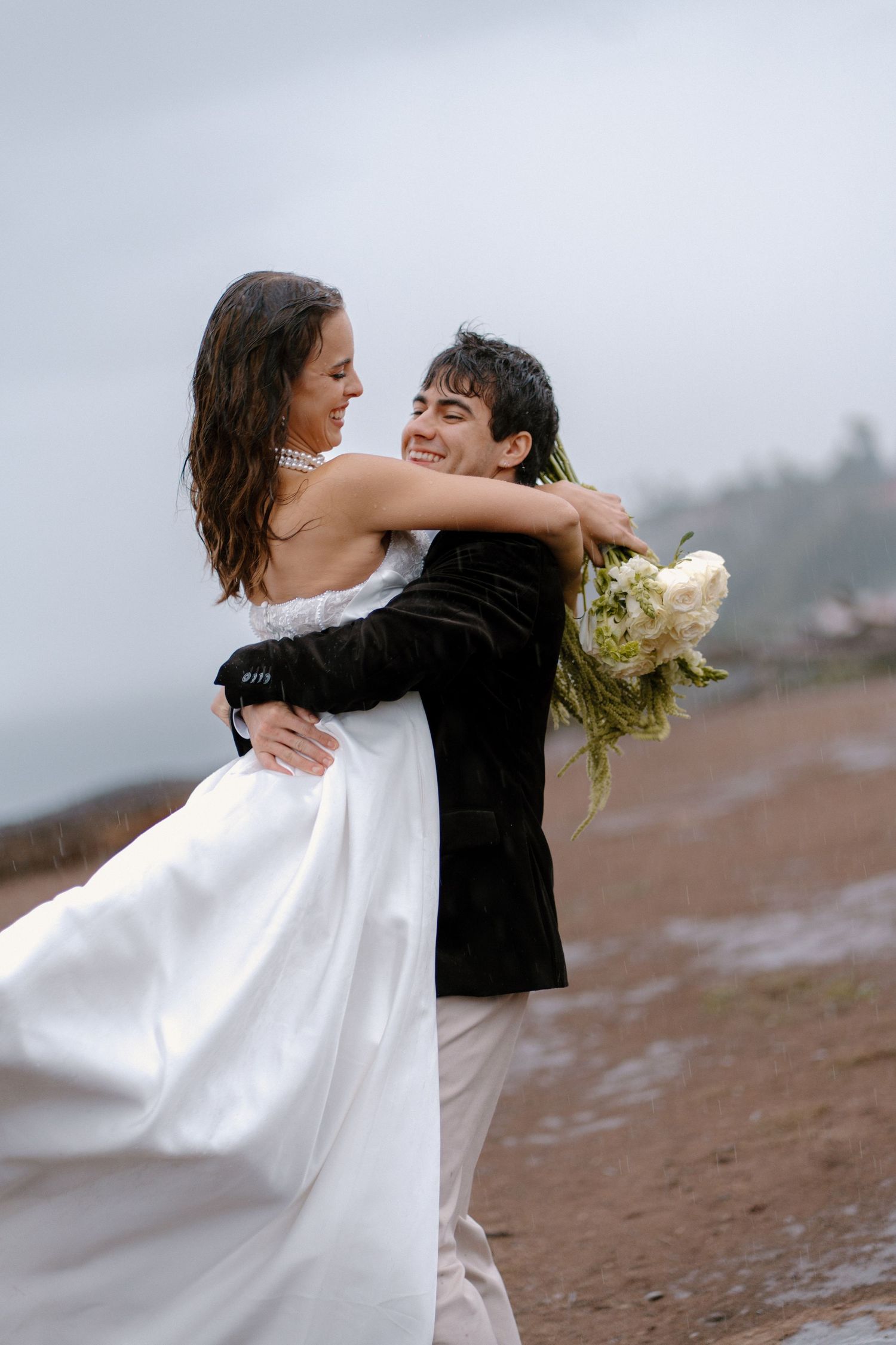 Groom lifts smiling bride in white gown on rainy beach, holding white bouquet.