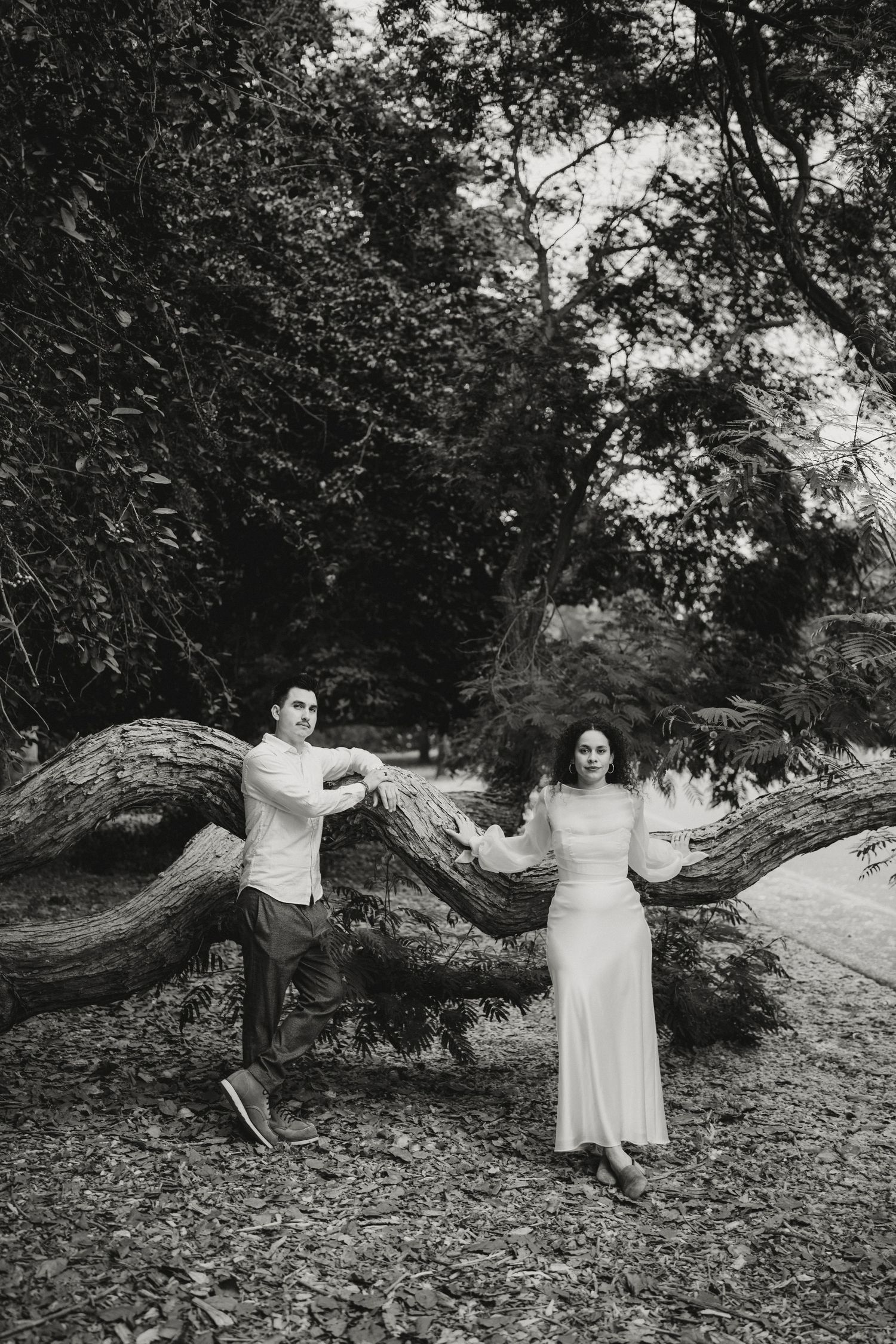 A couple poses beside a large fallen tree trunk in a moody black and white forest photo.