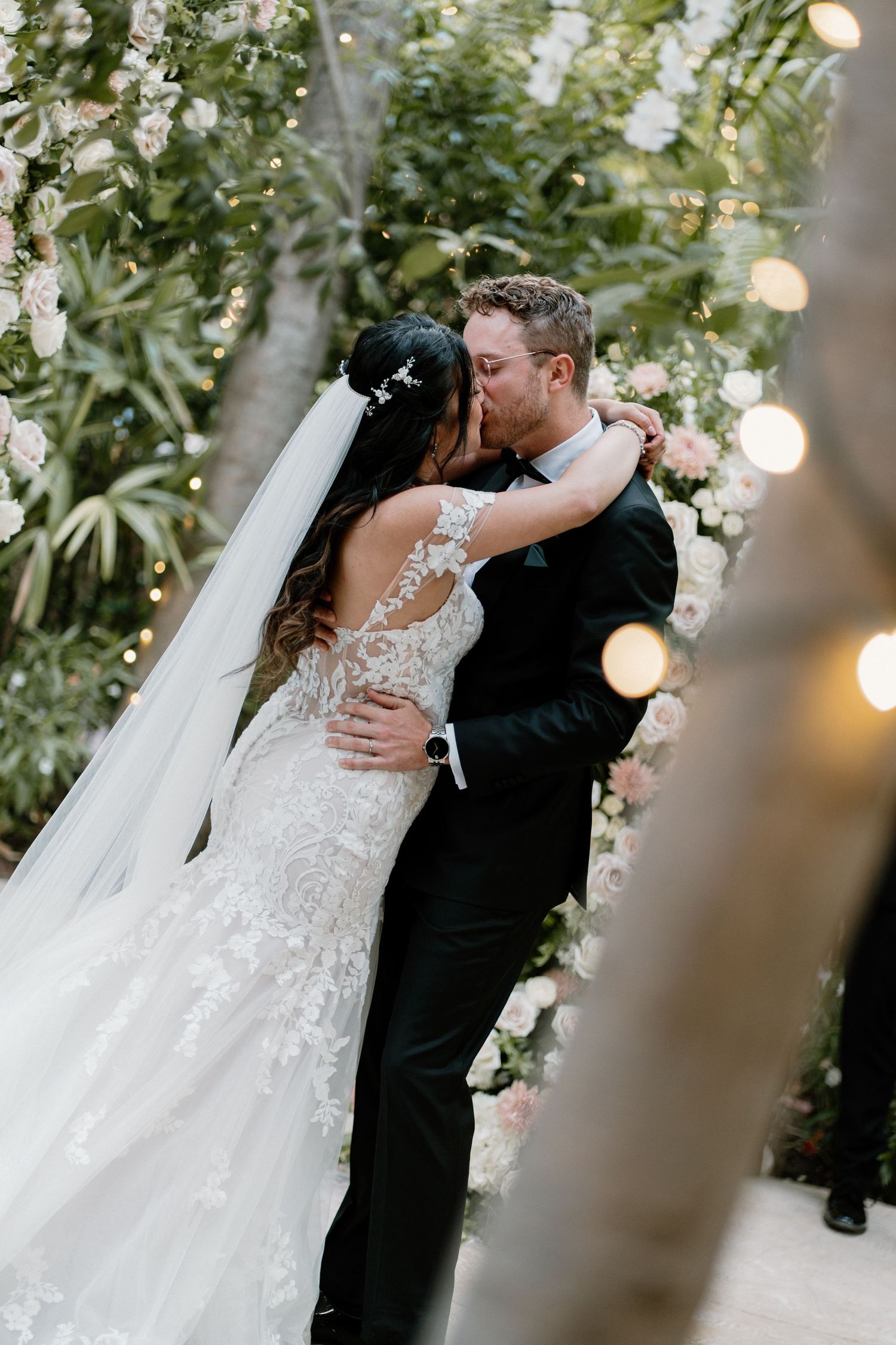 Bride and groom share their first kiss surrounded by floral arrangements and twinkling string lights.