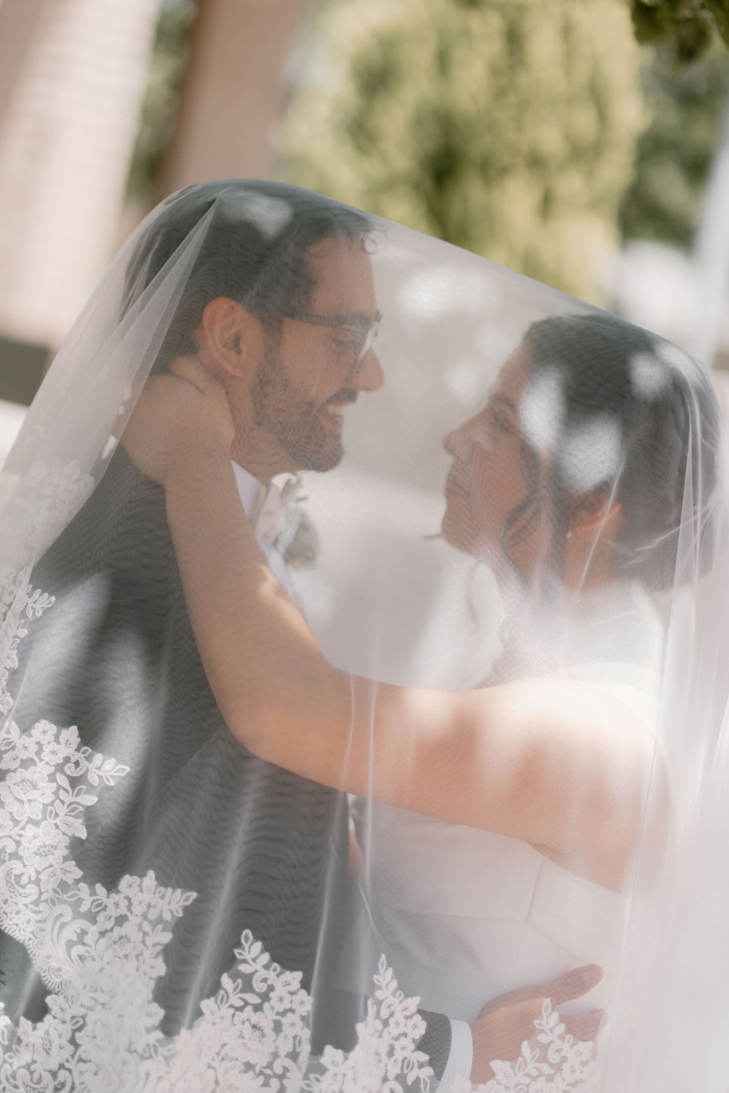 Bride and groom share a romantic moment under a lace wedding veil, gazing lovingly at each other outdoors.