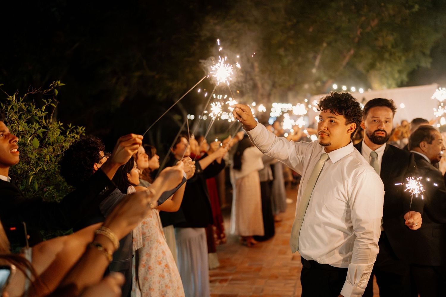 Wedding guests hold sparklers during nighttime sendoff celebration surrounded by trees and festive lighting.