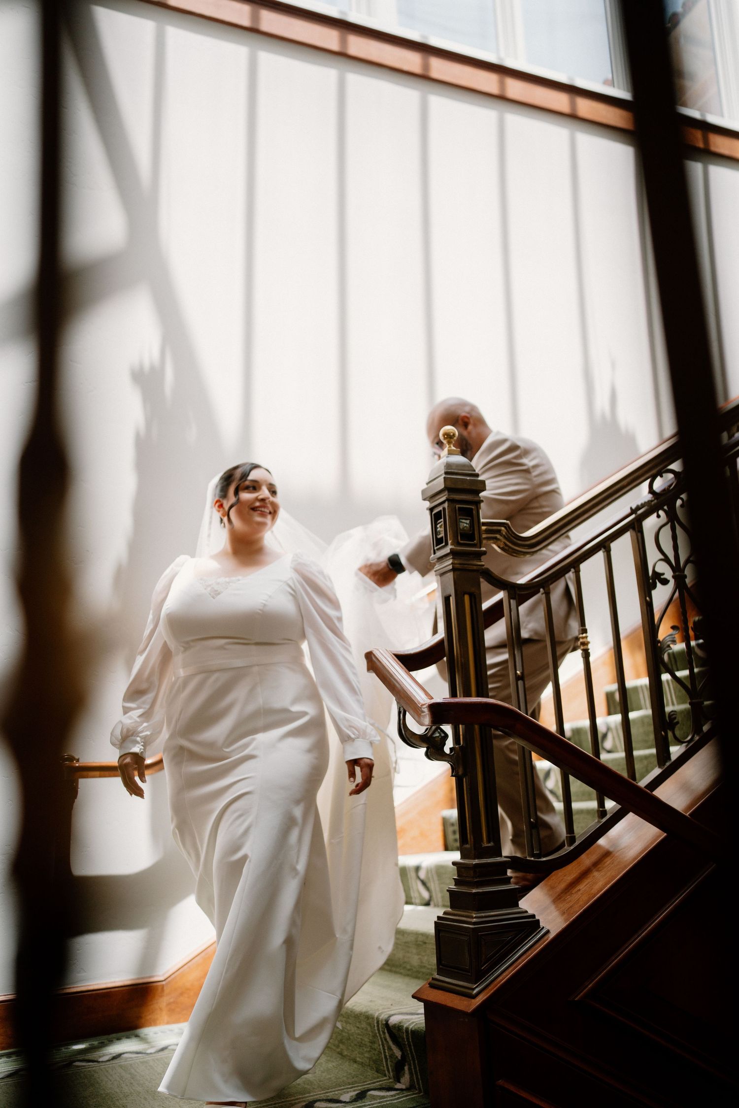 Bride in long white gown descending grand staircase with groom in sunlit wedding venue interior.