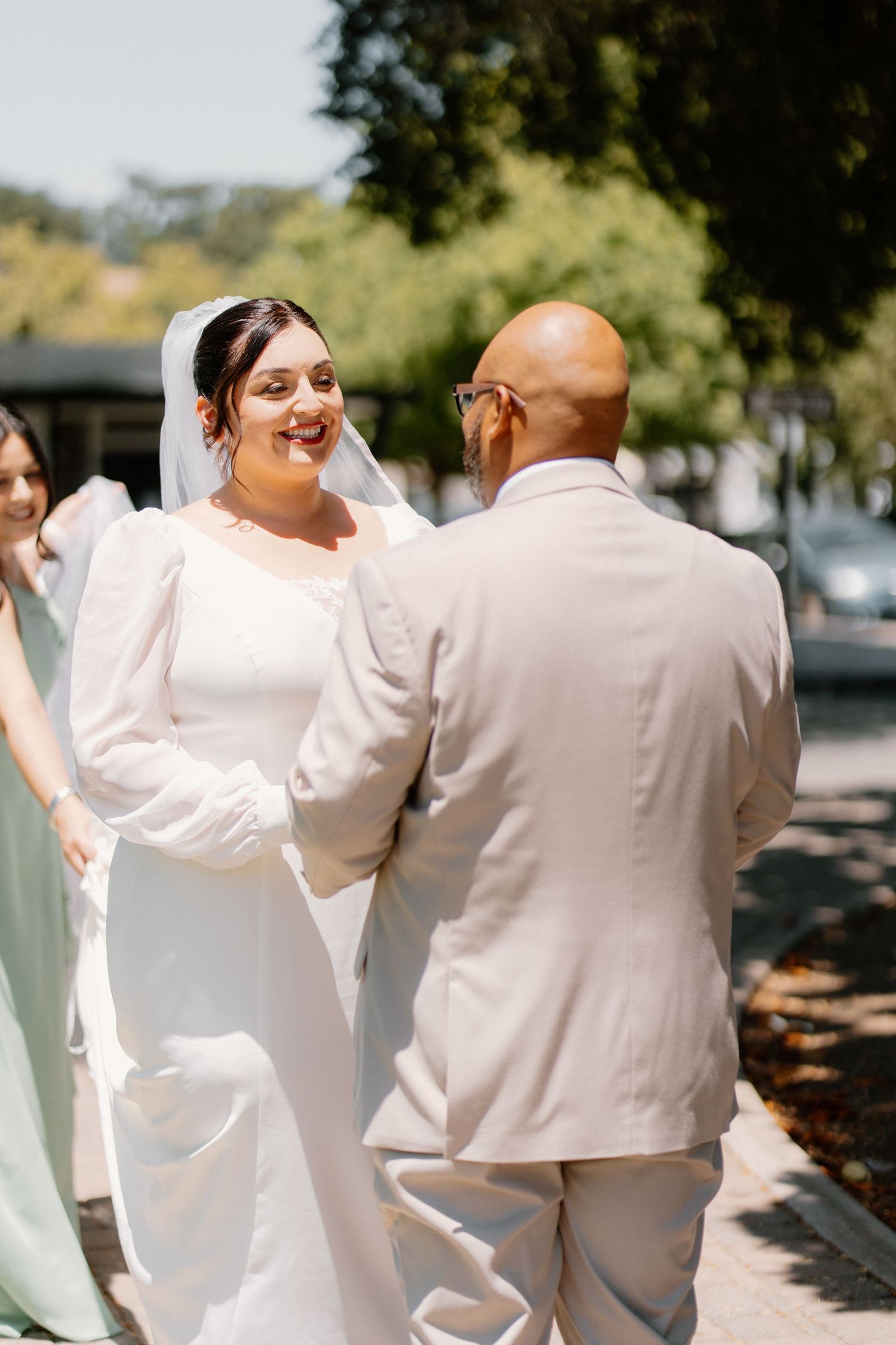 Smiling bride in white gown and veil holds hands with bald groom in tan suit outdoors on sunny day.