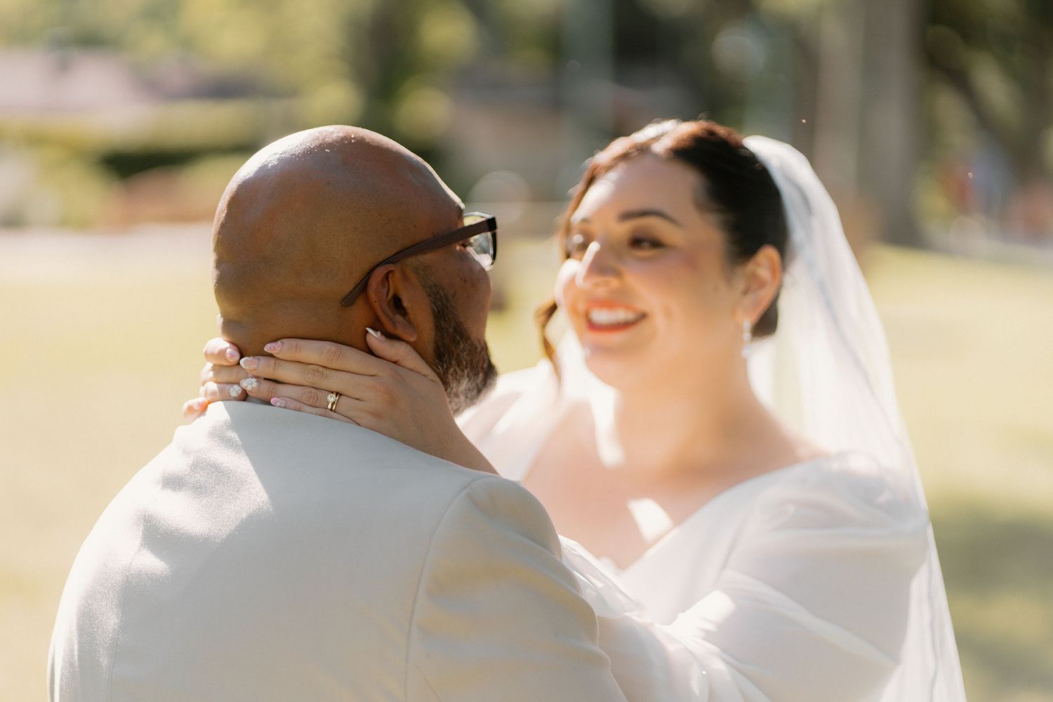 Bride in white veil smiles at groom outdoors during wedding ceremony, showing diamond ring on his face.