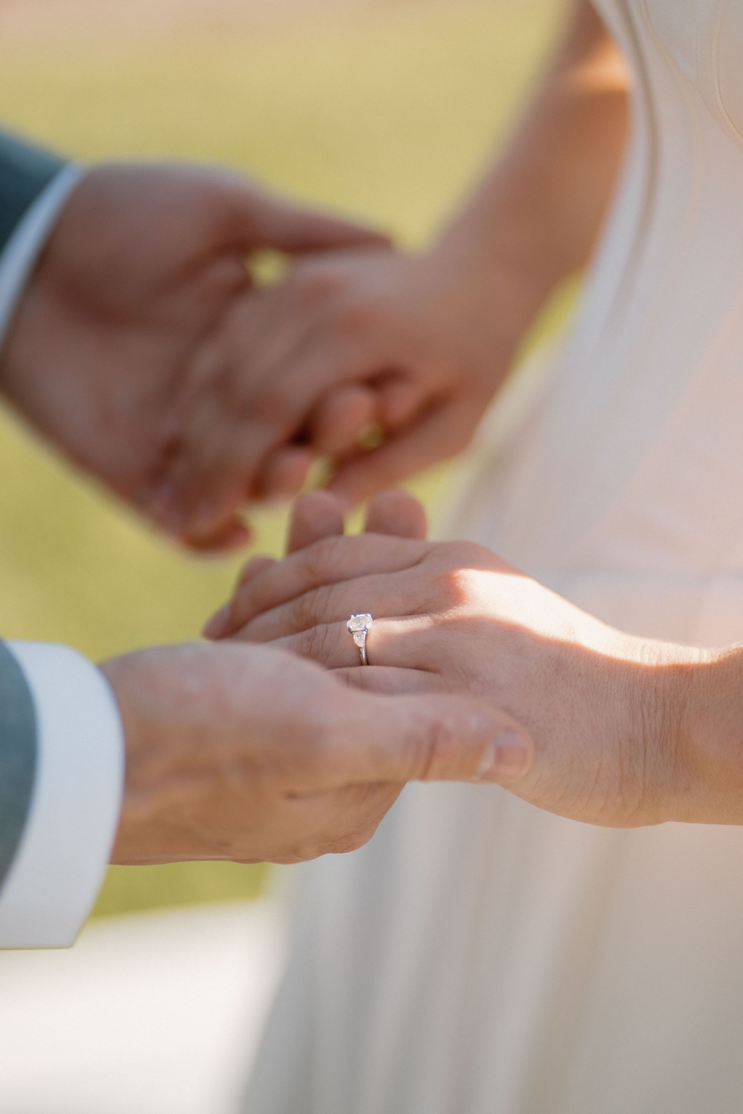 Groom placing a diamond solitaire engagement ring on brides finger during outdoor wedding ceremony.