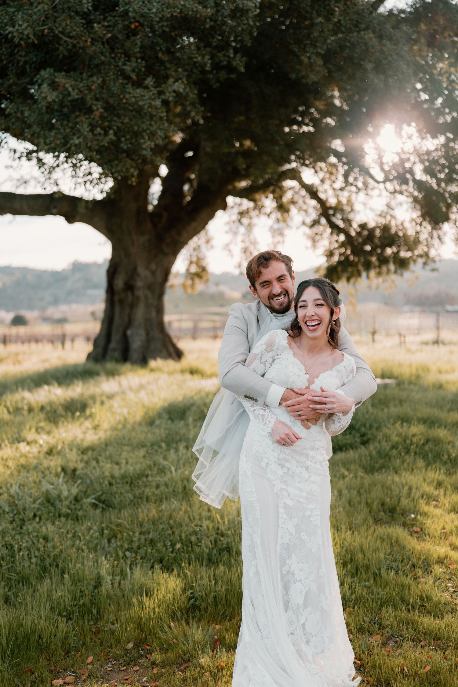 Groom hugging bride from behind under large oak tree in sunlit green meadow, both laughing.