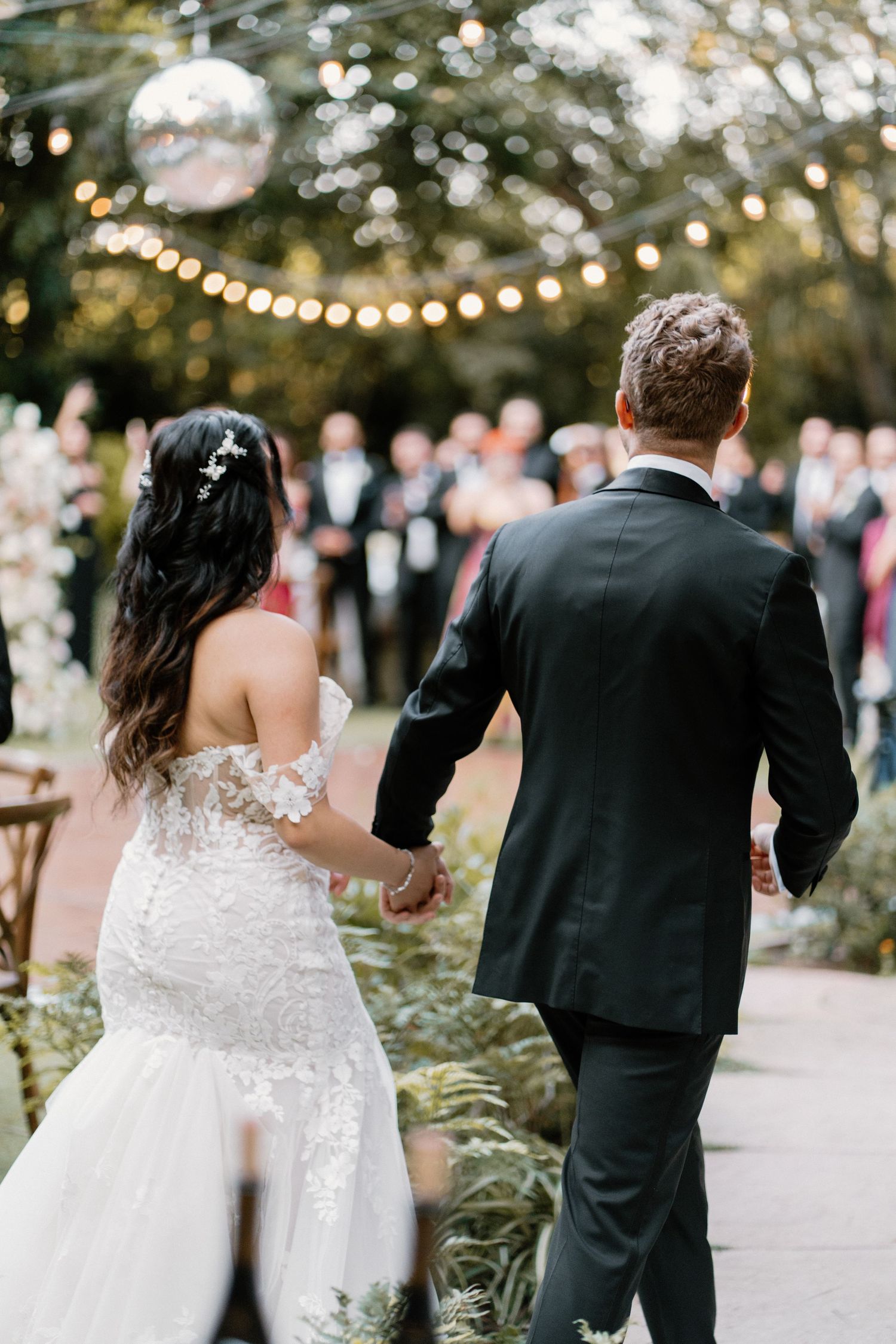 Bride and groom walk hand-in-hand toward guests under string lights and a disco ball outdoors.