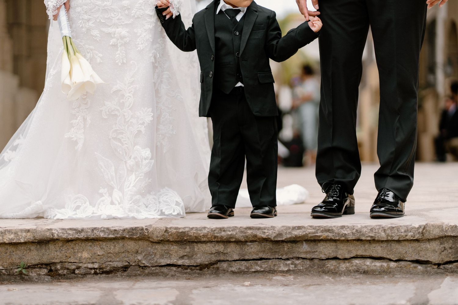 A young ring bearer in a black suit holds hands with the bride and groom at an outdoor wedding ceremony.