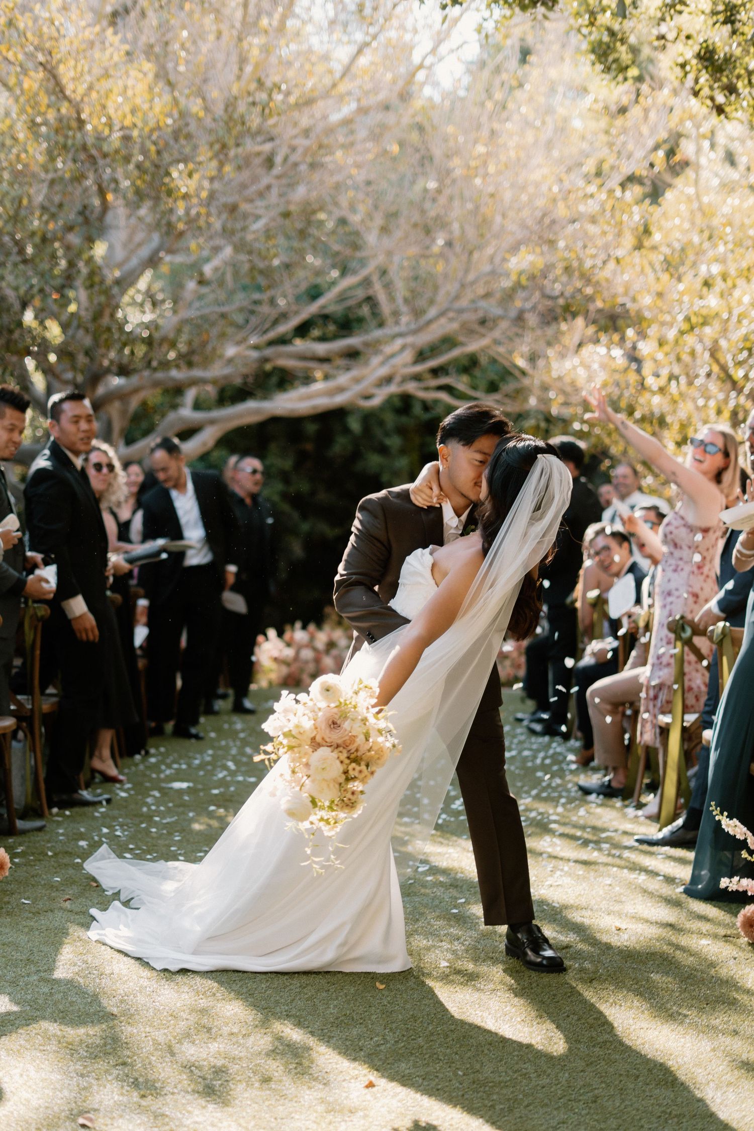 Groom dips bride for romantic kiss while walking down outdoor wedding aisle as guests cheer and toss petals.