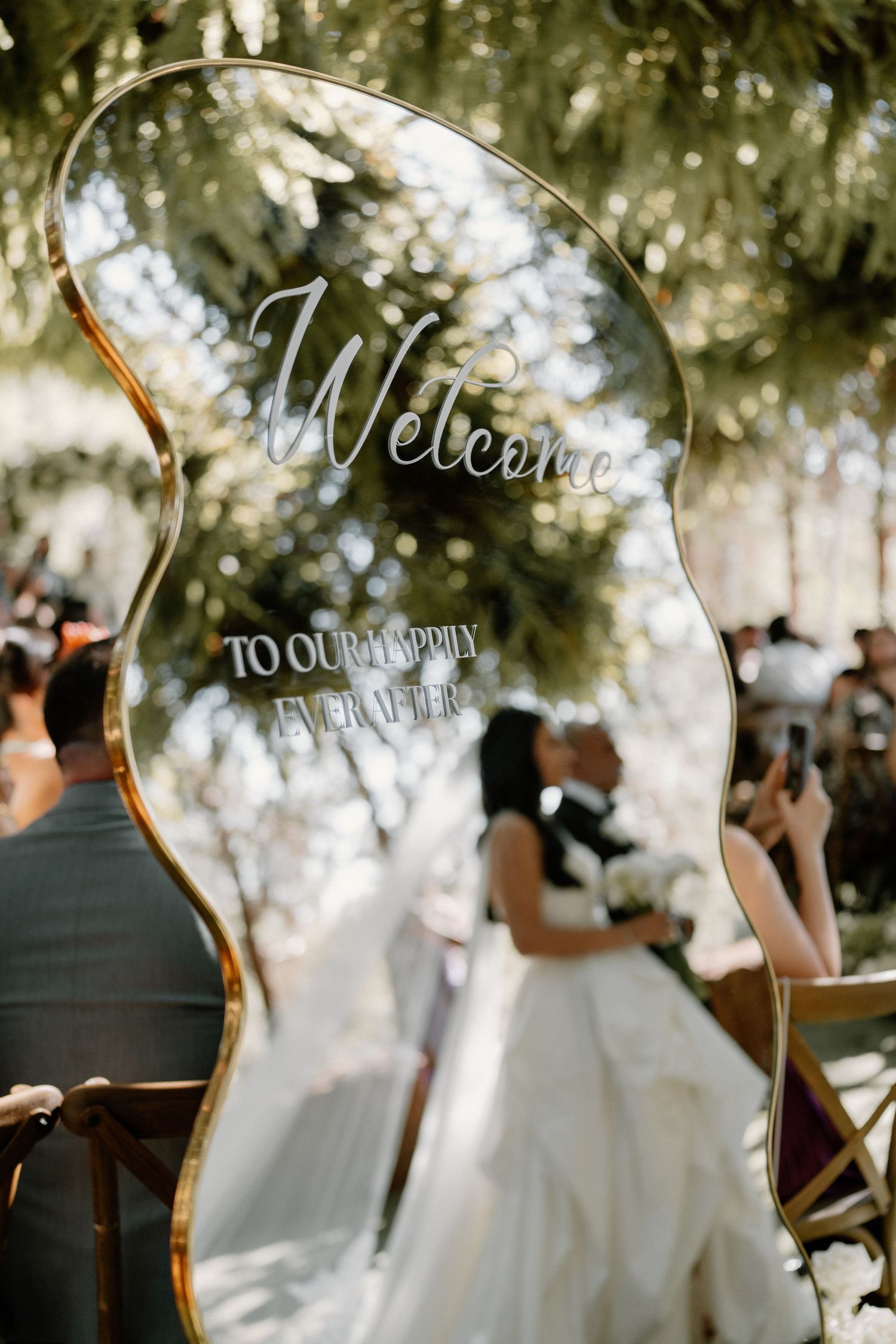 An elegant wavy gold-framed welcome sign reads Welcome To Our Happily Ever After at a wedding.