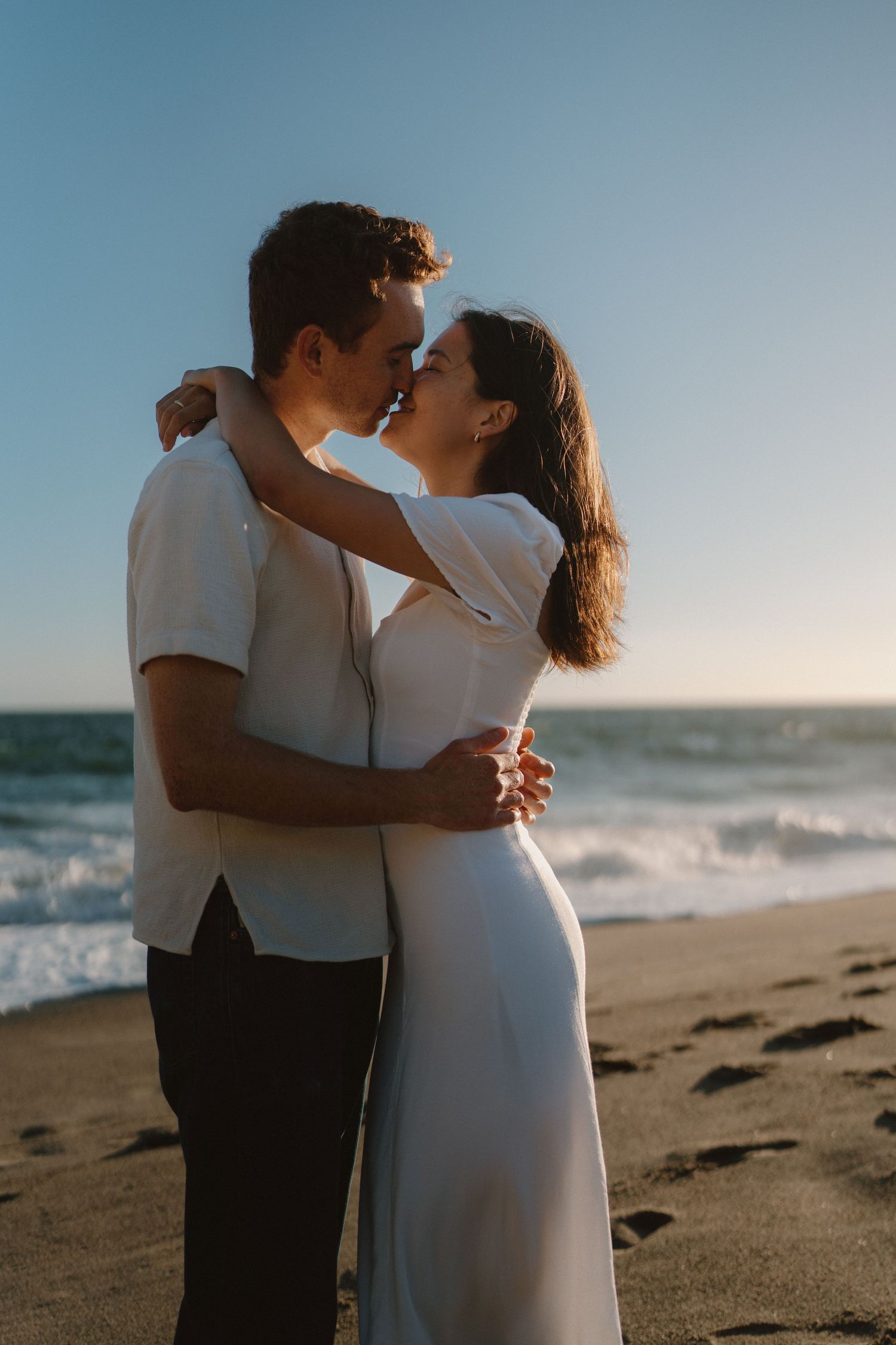 Couple sharing a romantic almost-kiss embrace on a sandy beach at sunset, woman in white dress, ocean waves behind them.