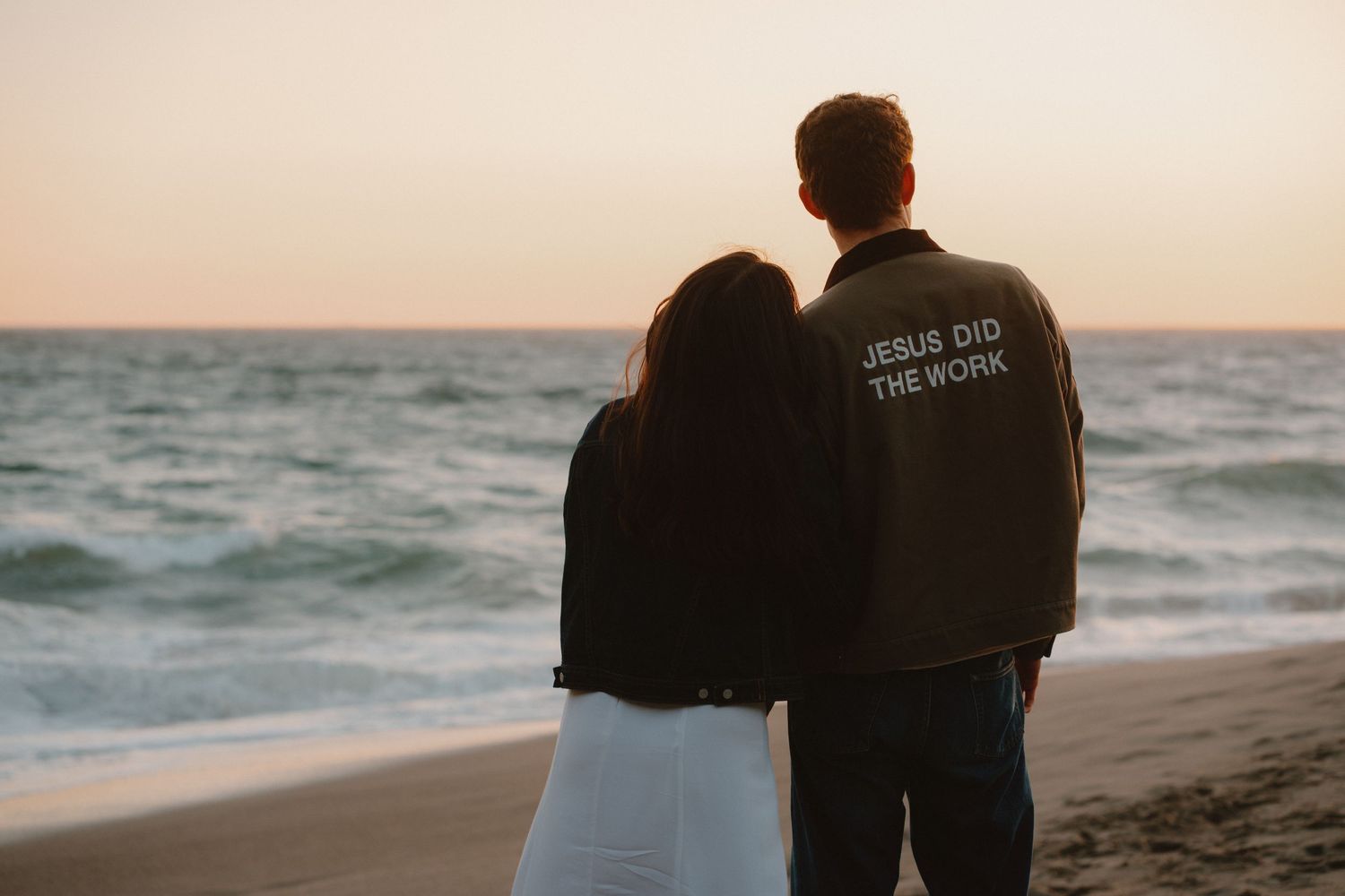 Couple standing together on a beach at sunset, man wearing a jacket reading Jesus Did The Work.