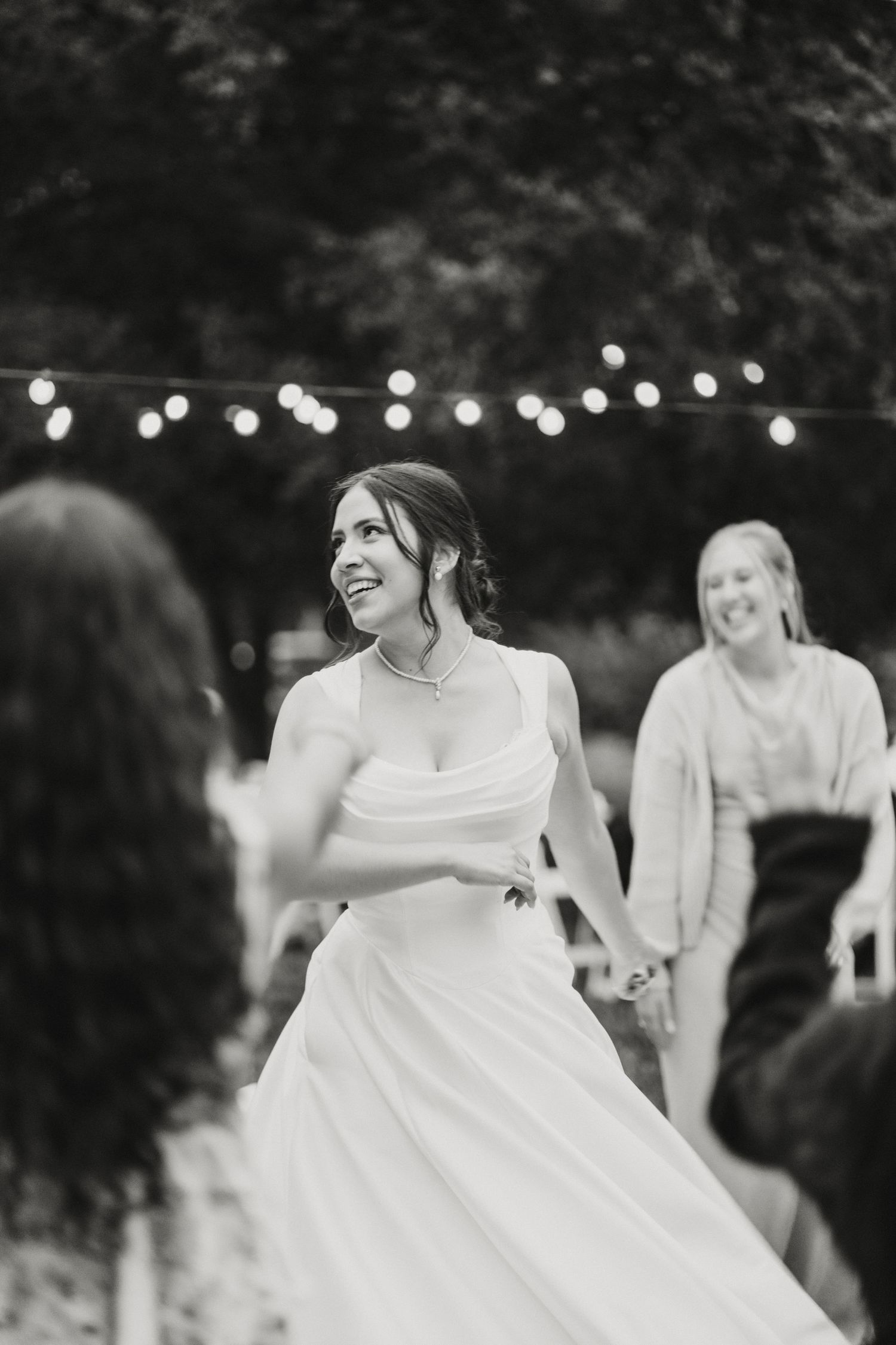 Black and white photo of a joyful bride dancing with guests at an outdoor wedding reception.