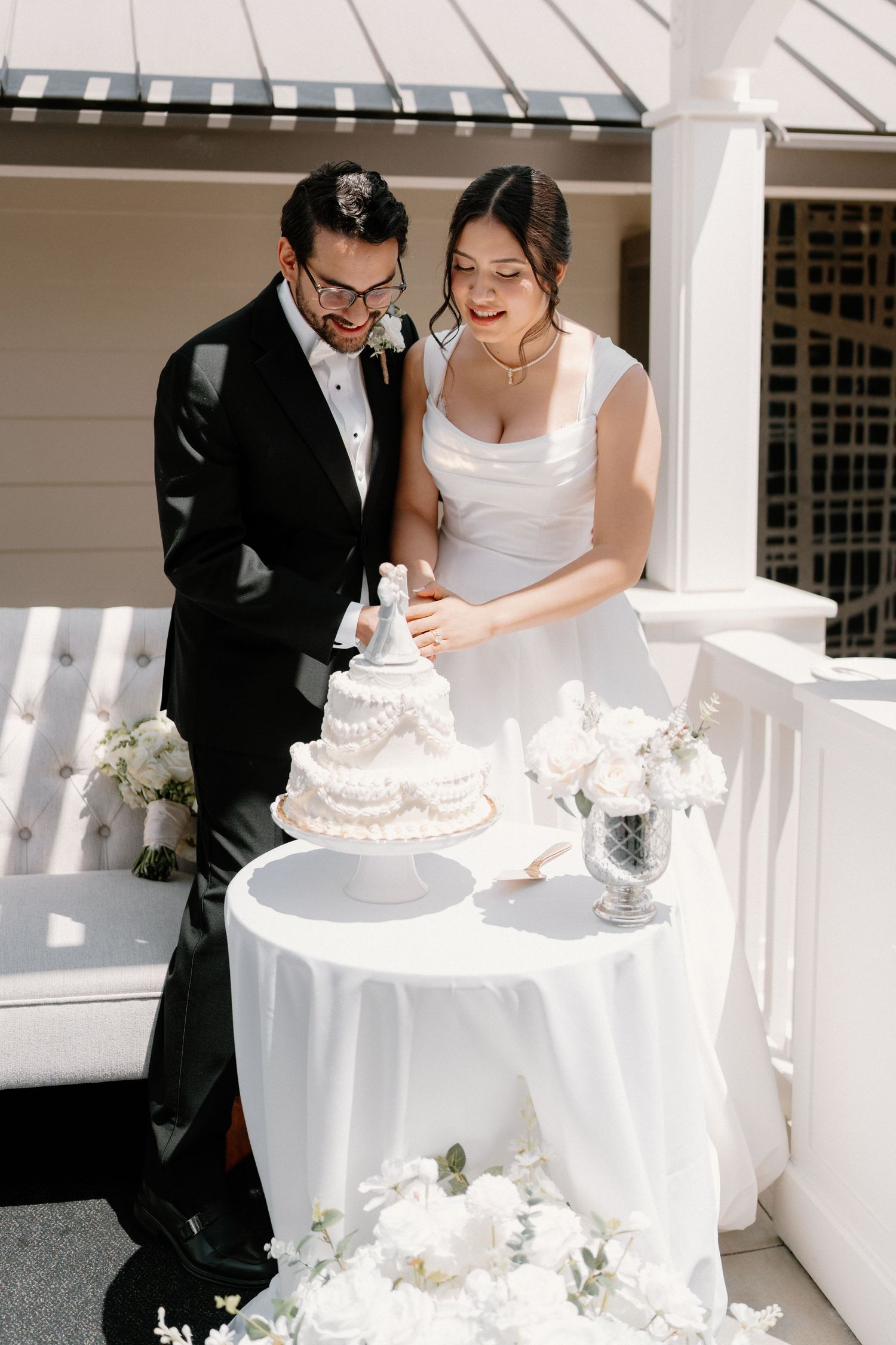Bride and groom cut elegant white tiered wedding cake on sunlit outdoor venue porch decorated with white flowers.