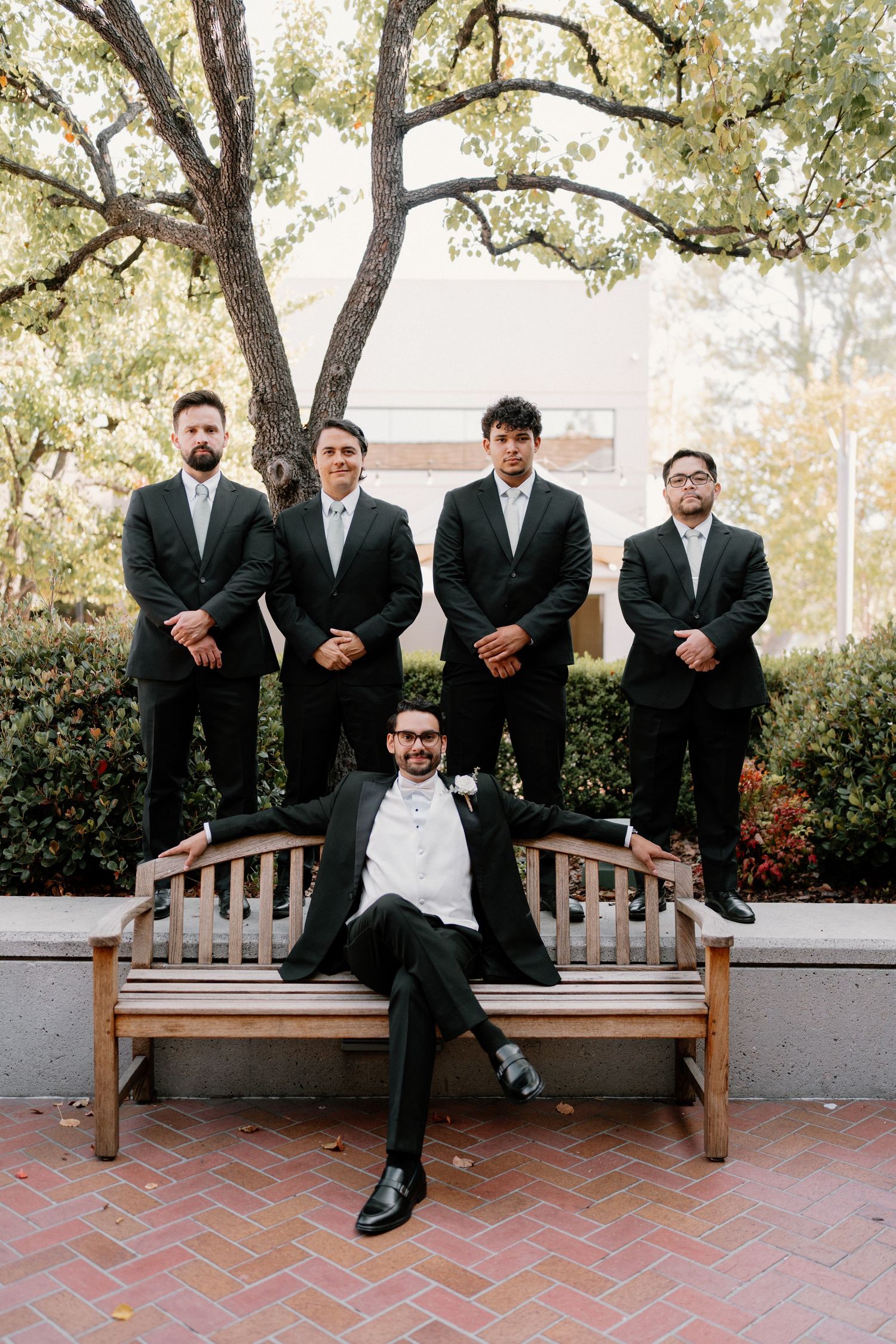 Groom poses on bench with four groomsmen standing behind him in dark suits outdoors.