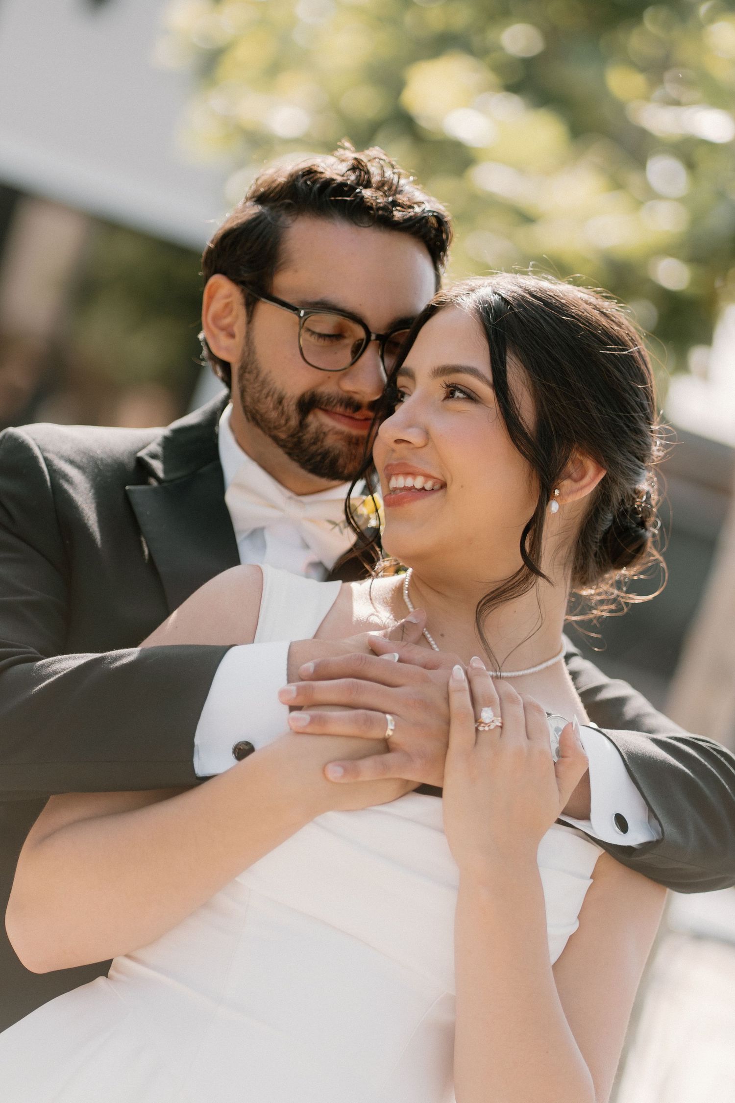 Bride and groom embrace outdoors, she smiles wearing a white gown and diamond ring, he wears a dark suit with glasses.