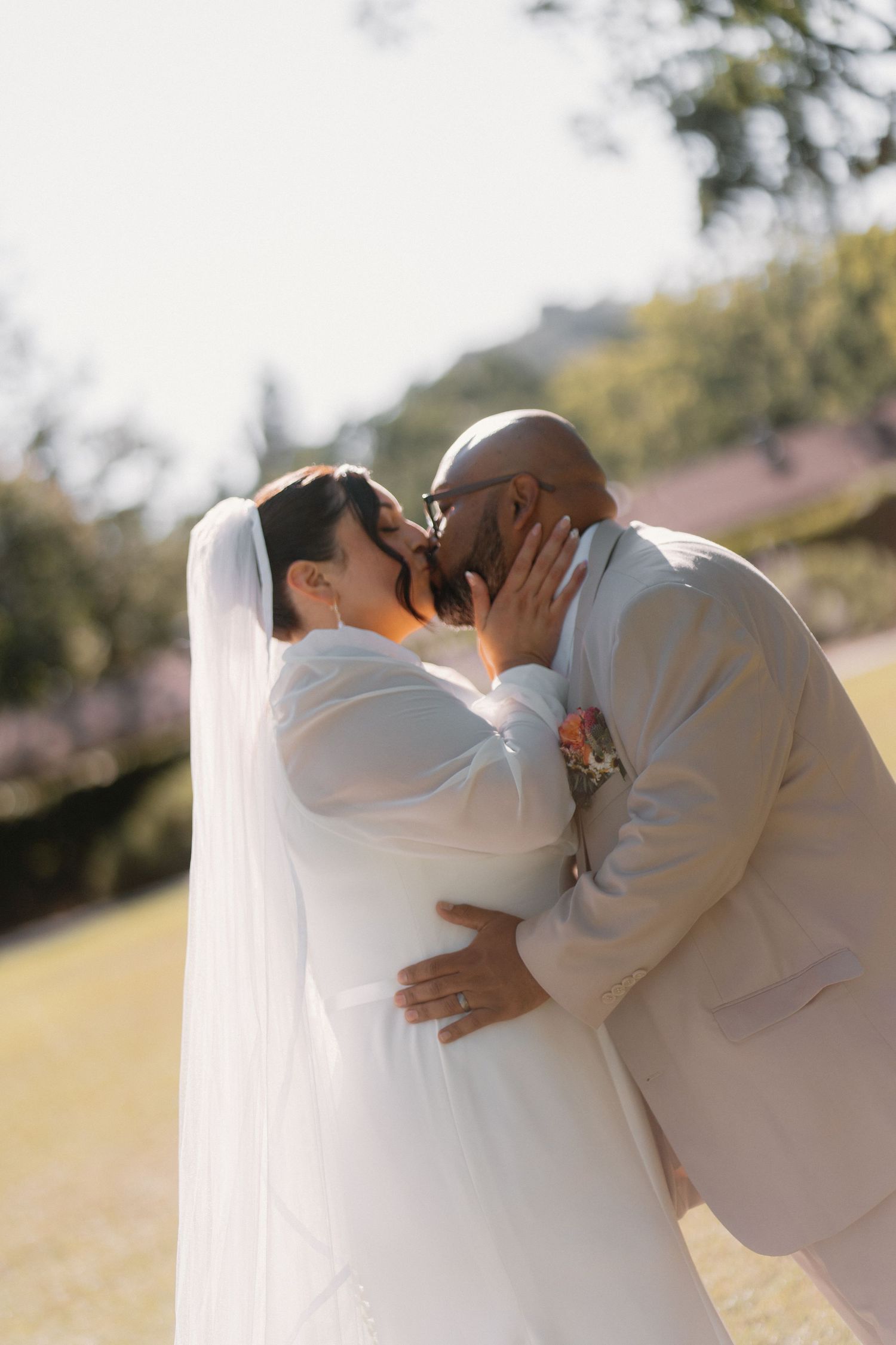 Bride in white gown and veil shares a romantic kiss with groom in tan suit outdoors.