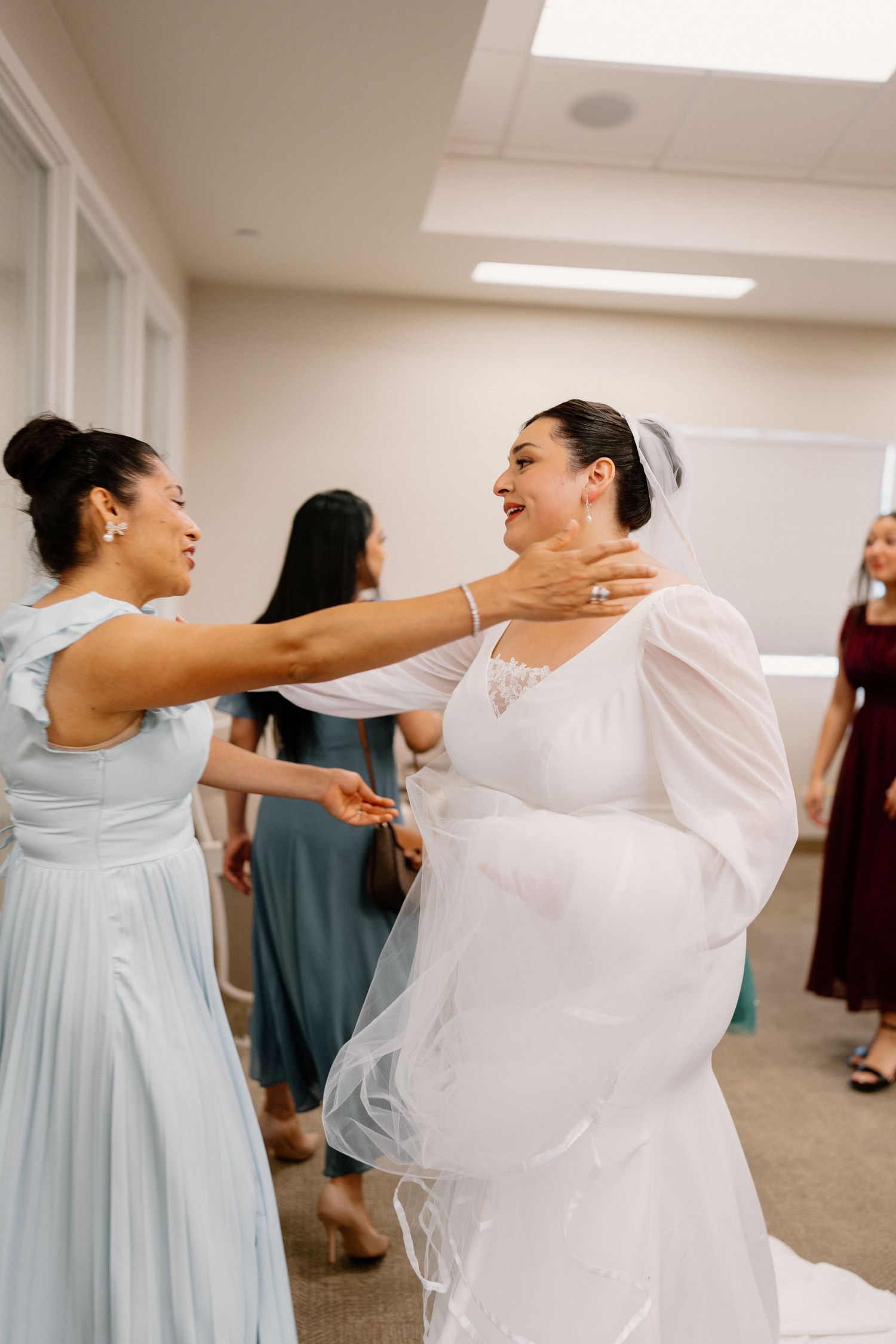Bridesmaid in light blue dress helps bride in white gown prepare before wedding ceremony.