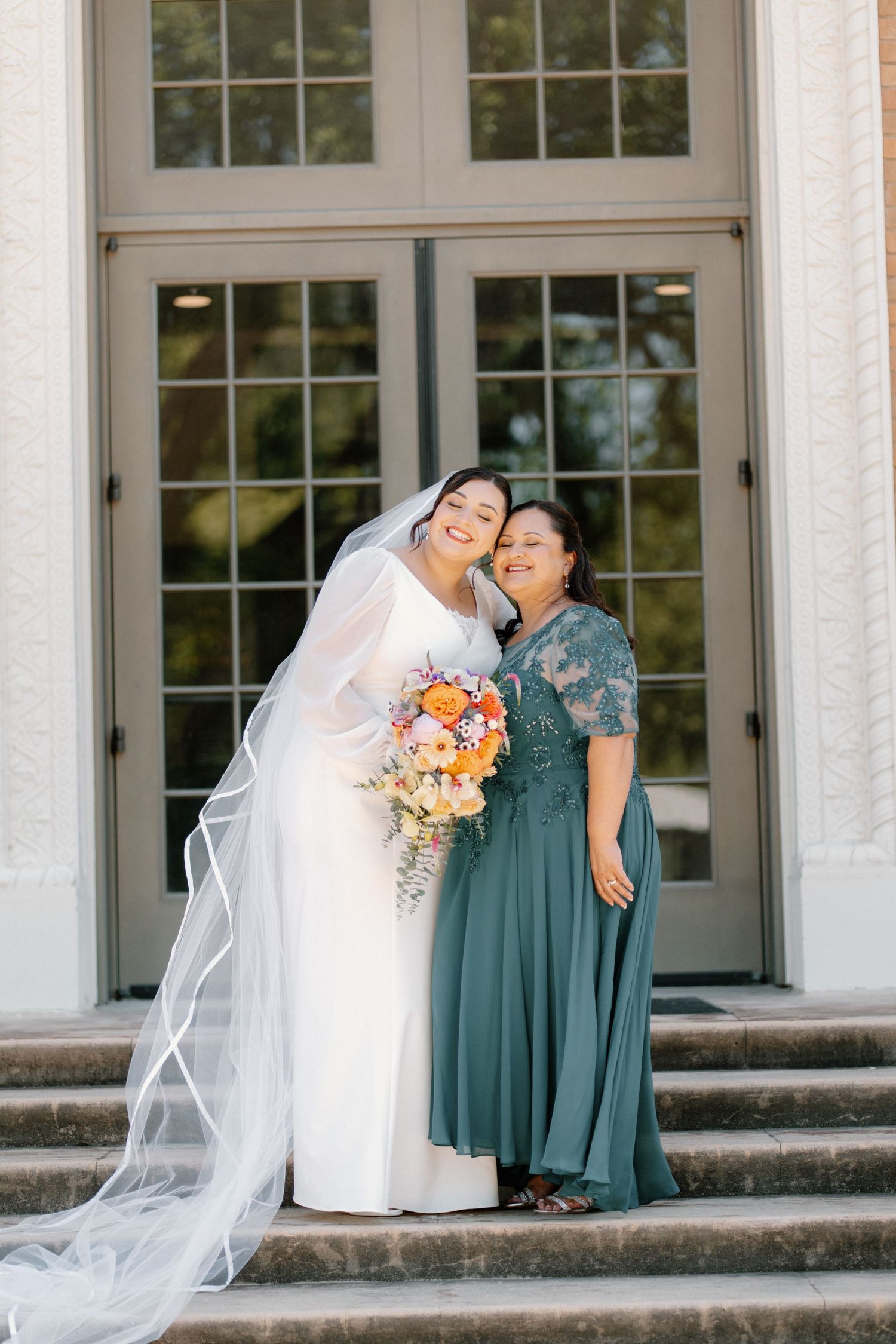 Bride in white gown and veil poses with woman in teal dress holding bouquet on steps.