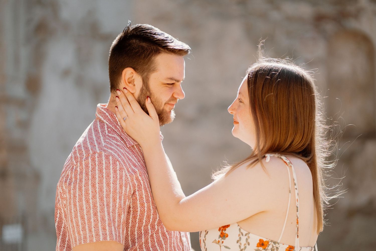 Couple sharing a tender moment outdoors, woman gently touching man's face, warm golden sunlight in background.