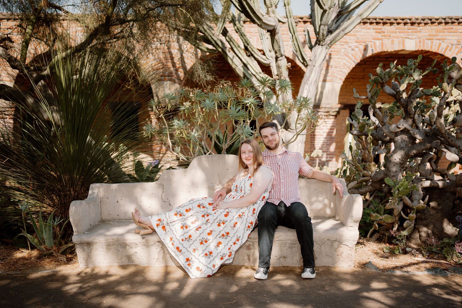 Couple sitting on white outdoor sofa surrounded by cacti and tropical plants in a sunny desert garden setting.