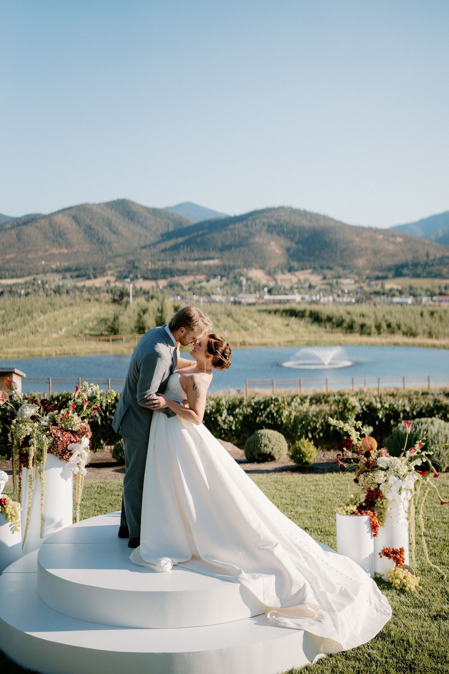 Bride and groom share a kiss on a circular platform with mountain lake views and floral arrangements at an outdoor wedding.