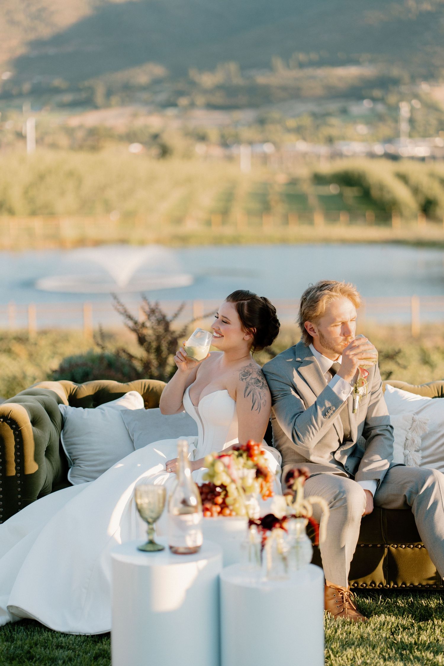 Bride and groom toast with wine glasses on an outdoor velvet sofa with a lake fountain backdrop.
