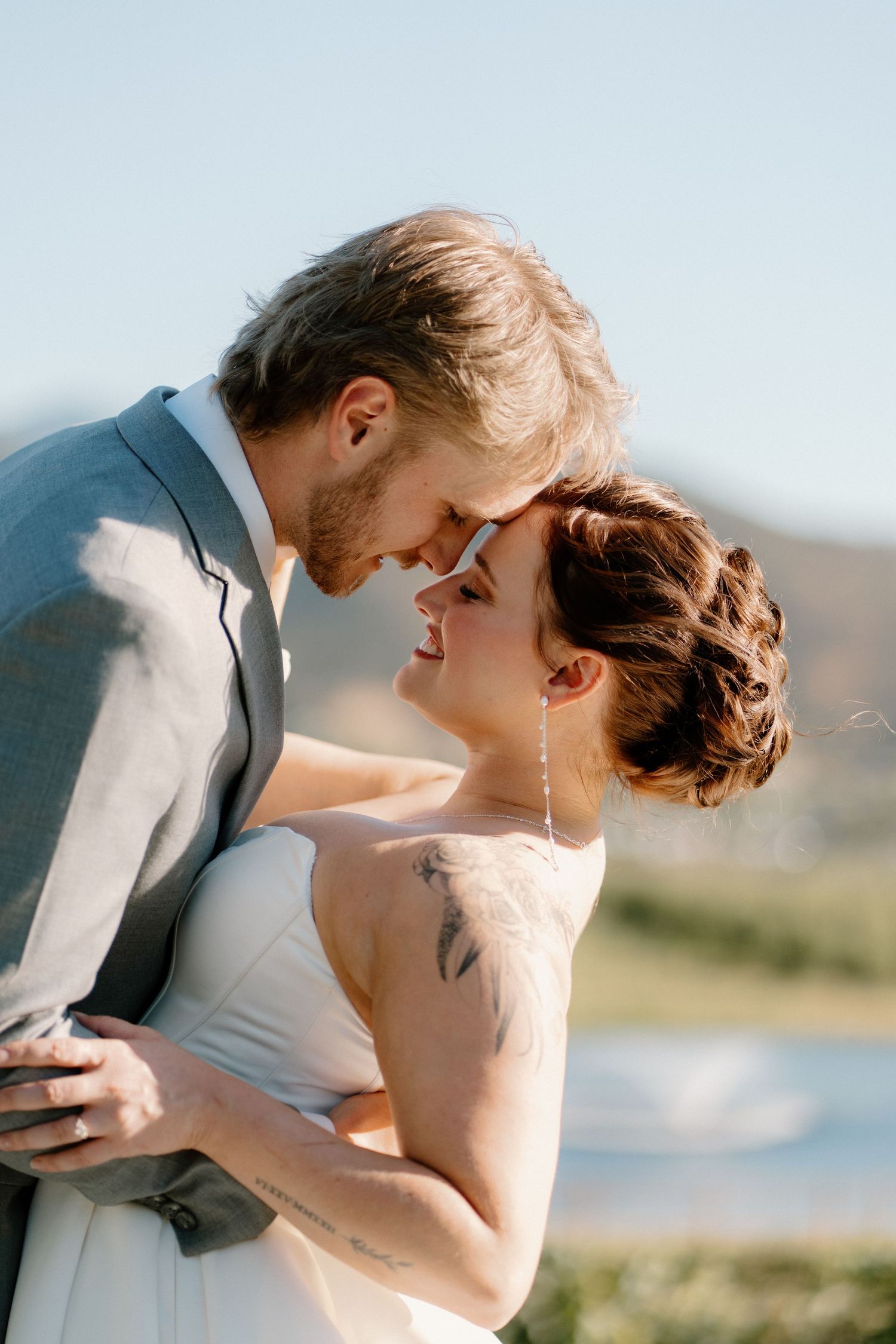 Bride and groom share romantic moment touching foreheads outdoors with scenic mountain view.