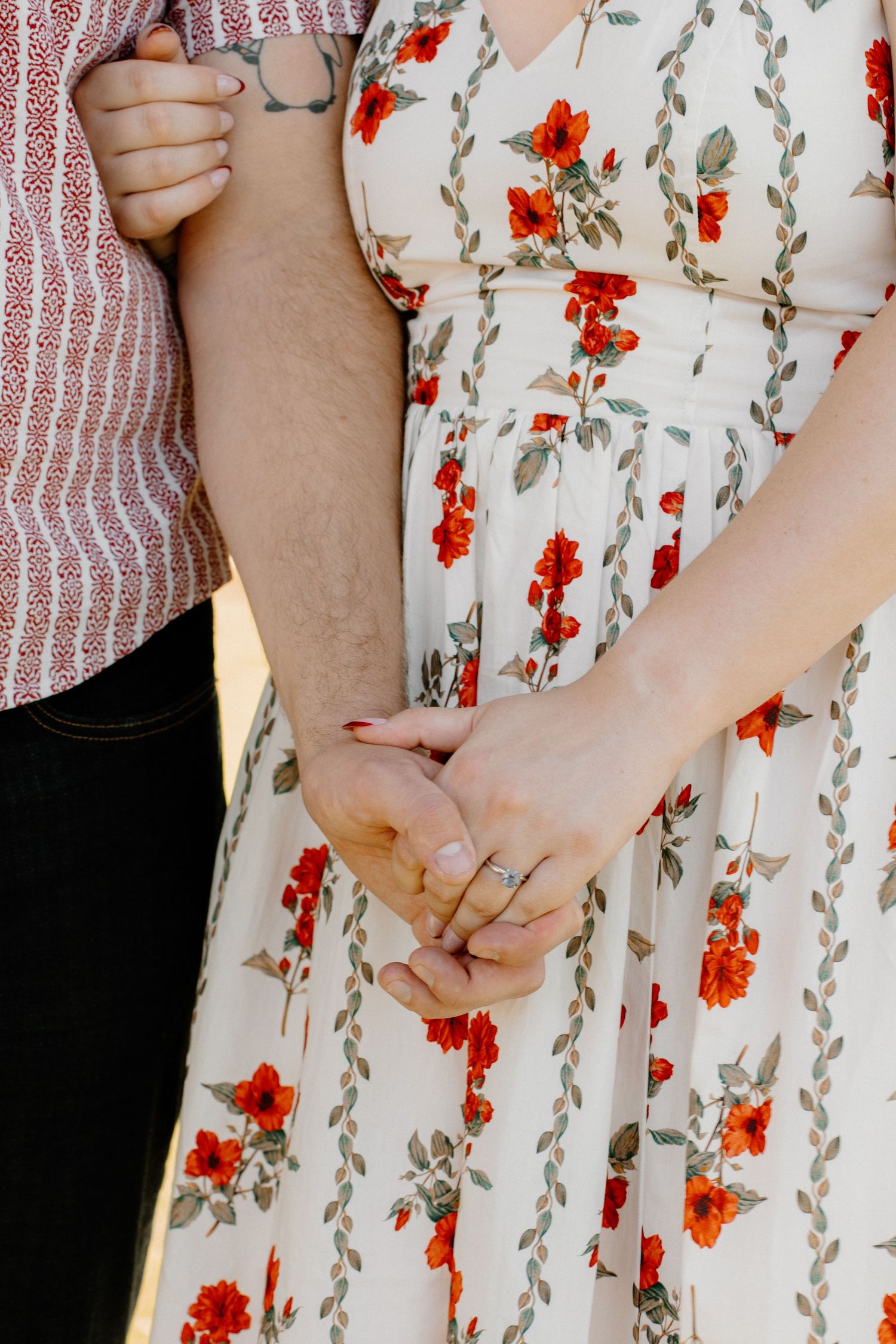 Couple holding hands, woman wearing white floral dress with red flowers, man in striped shirt, engagement ring visible.