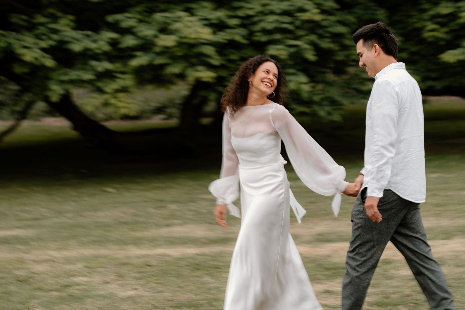 Smiling couple walking hand in hand through a green park during an outdoor engagement session.