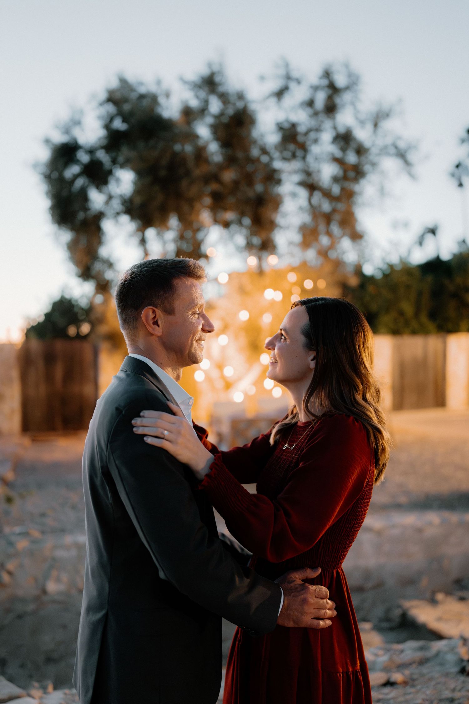 Couple embracing and smiling at each other with bokeh string lights at twilight.