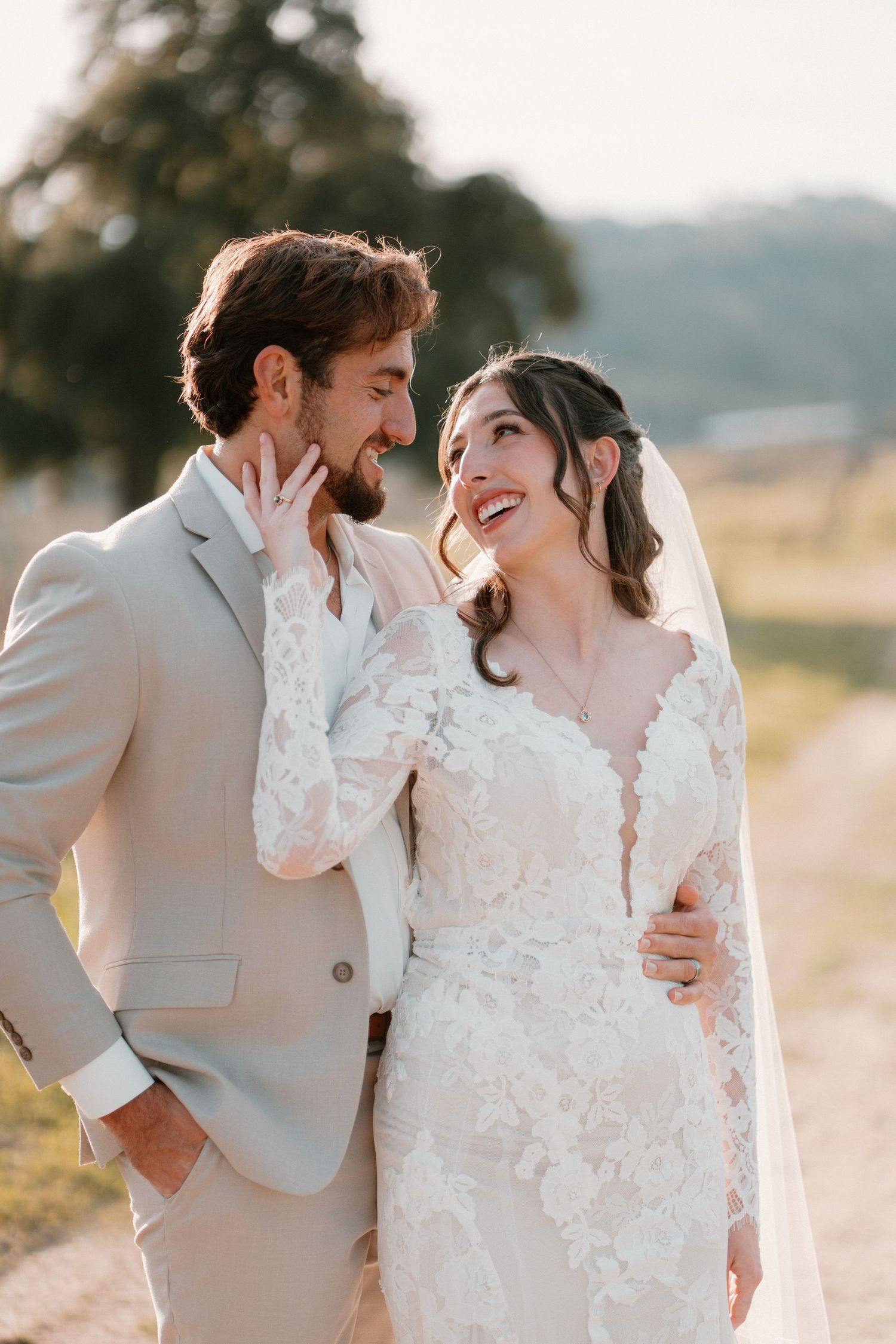 Bride and groom share a romantic moment laughing together outdoors in golden hour light, she in lace gown, he in tan suit.