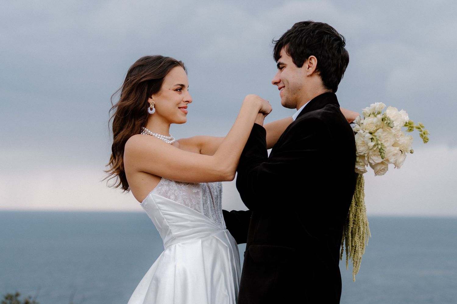 Bride and groom share a joyful moment by the ocean, she adjusts his tuxedo lapel while holding white flowers.